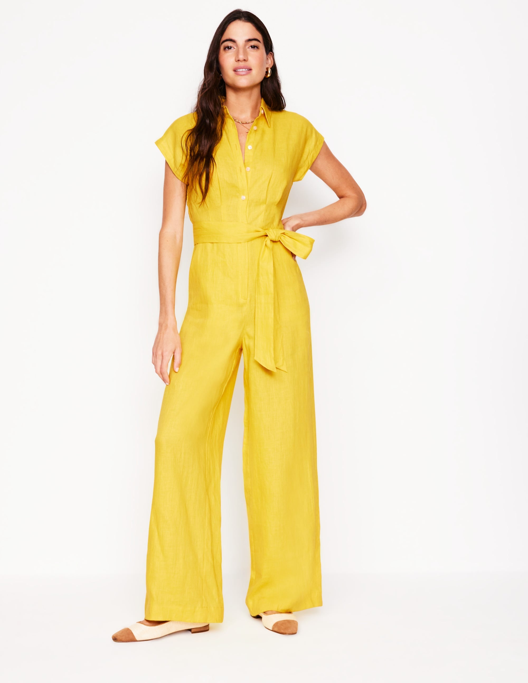 A woman wearing a bright yellow linen jumpsuit with short sleeves, a tie belt, and wide-leg pants.
