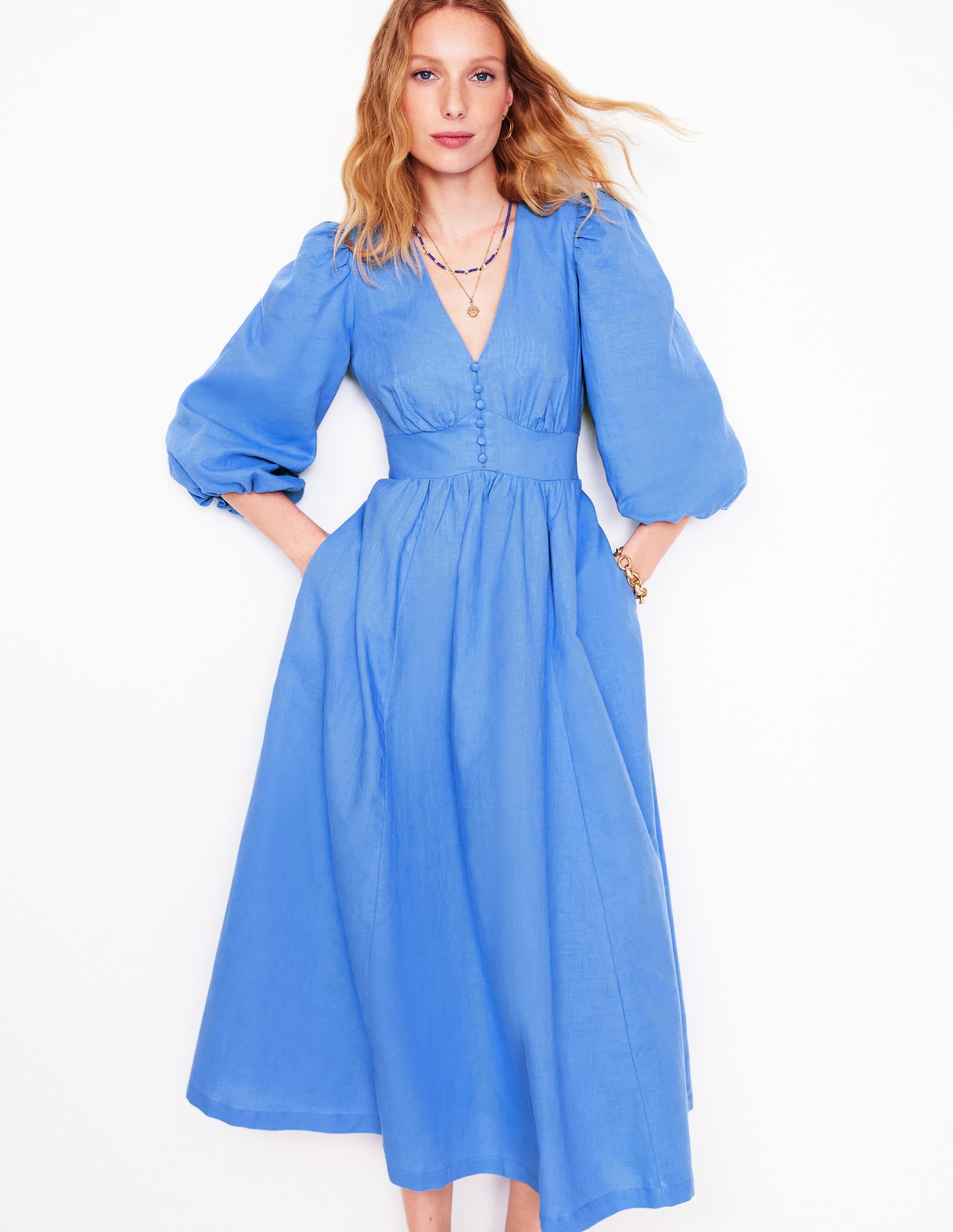 A woman wearing a blue maxi dress with a deep V-neckline, blouson bodice and sleeves, and side seam pockets.