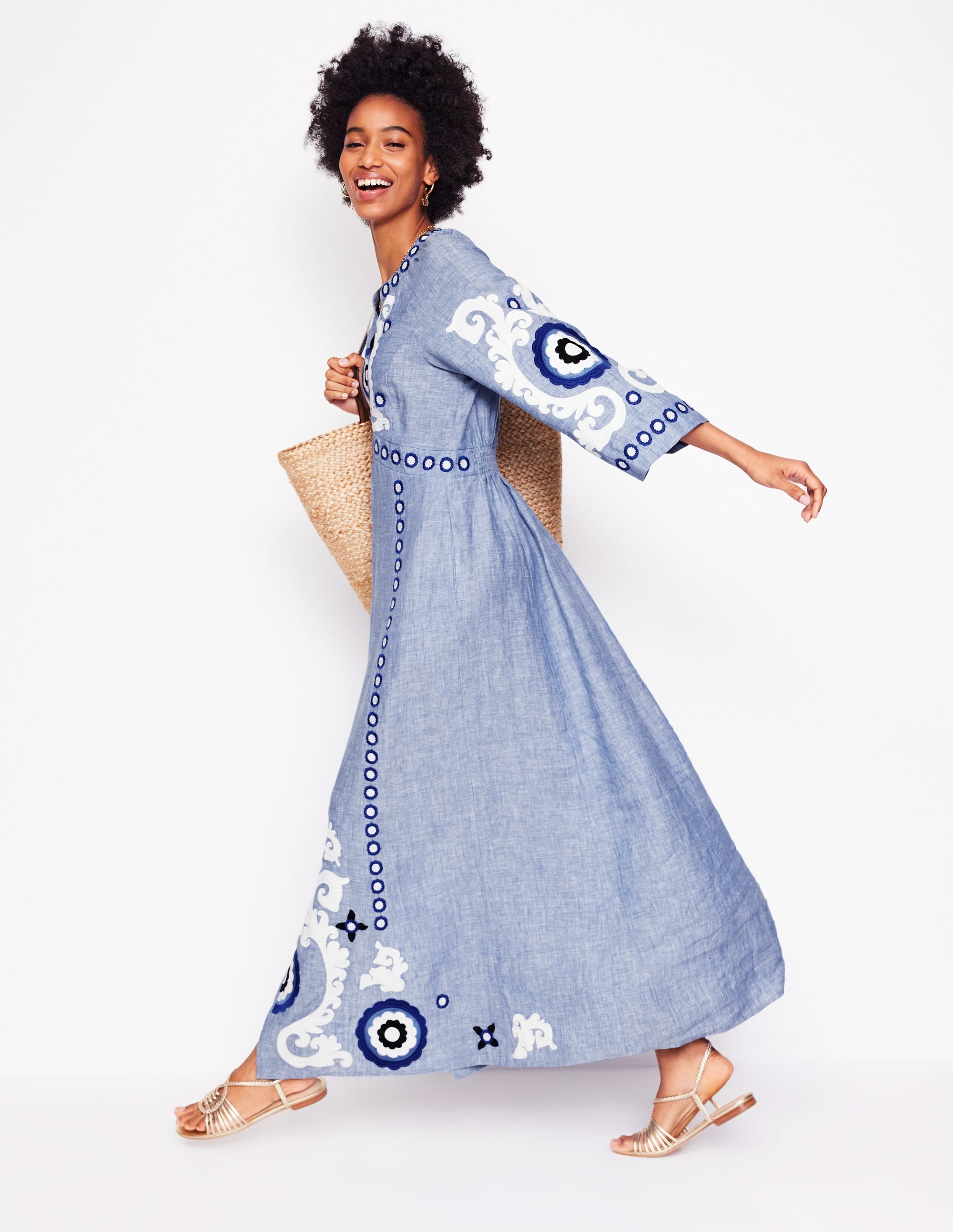 A woman wearing a light blue linen embroidered maxi dress with white and dark blue embroidery, holding a straw bag, and wearing sandals.