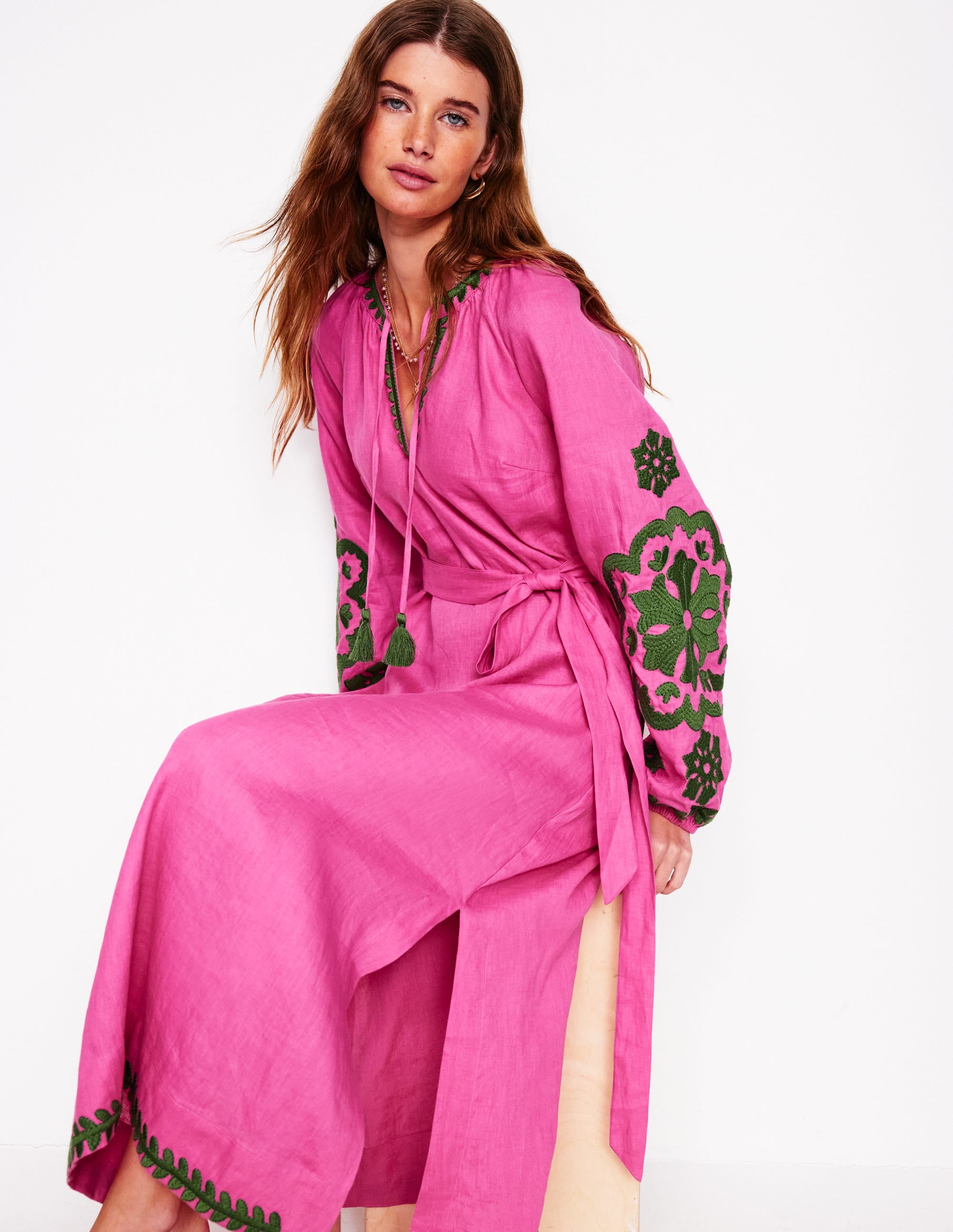 A woman wearing a bright pink linen dress with green embroidered floral designs on the sleeves and hem, with tassel details at the neckline.