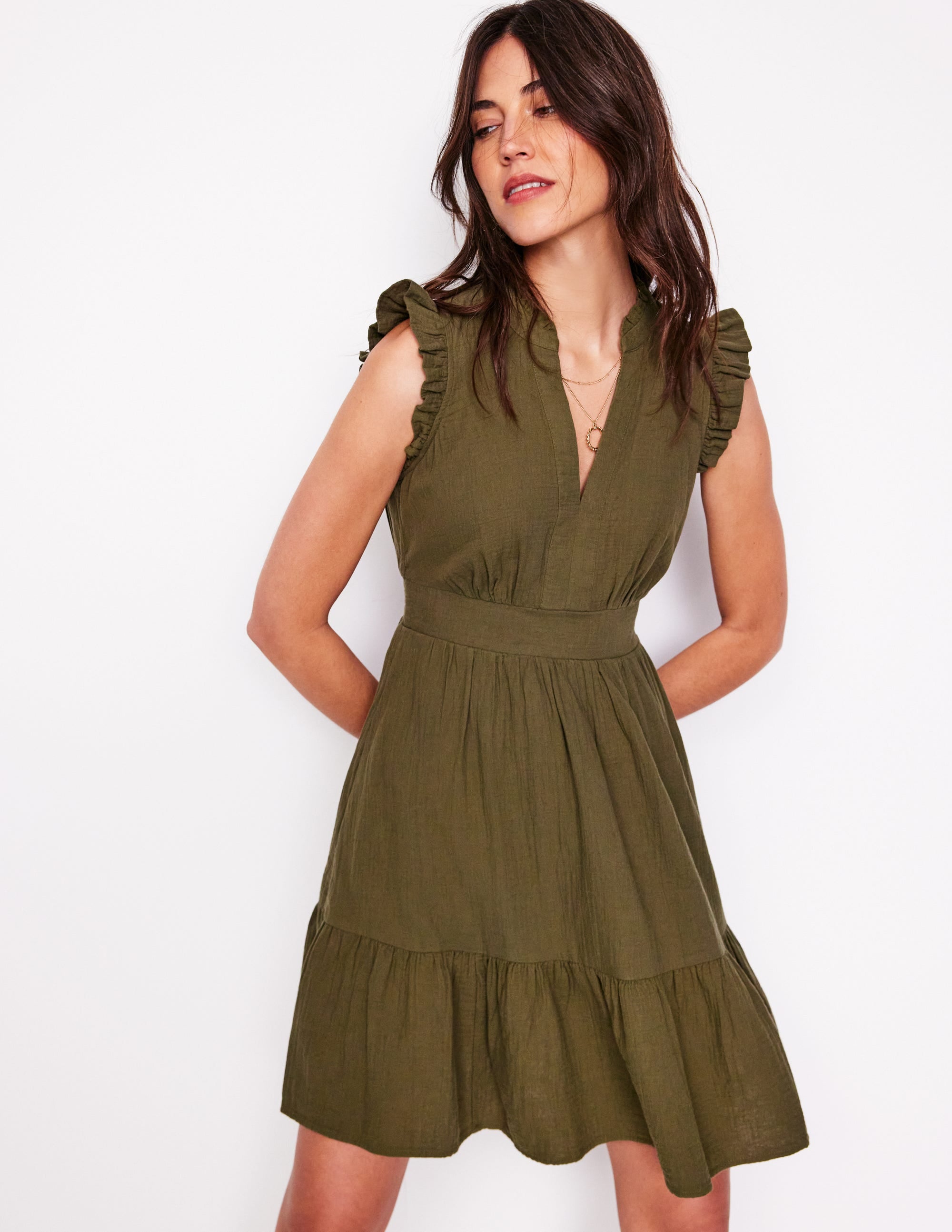 A woman wearing an olive green short dress with ruffled shoulder details, a V-neckline, and a ruffled hem, standing against a white background.
