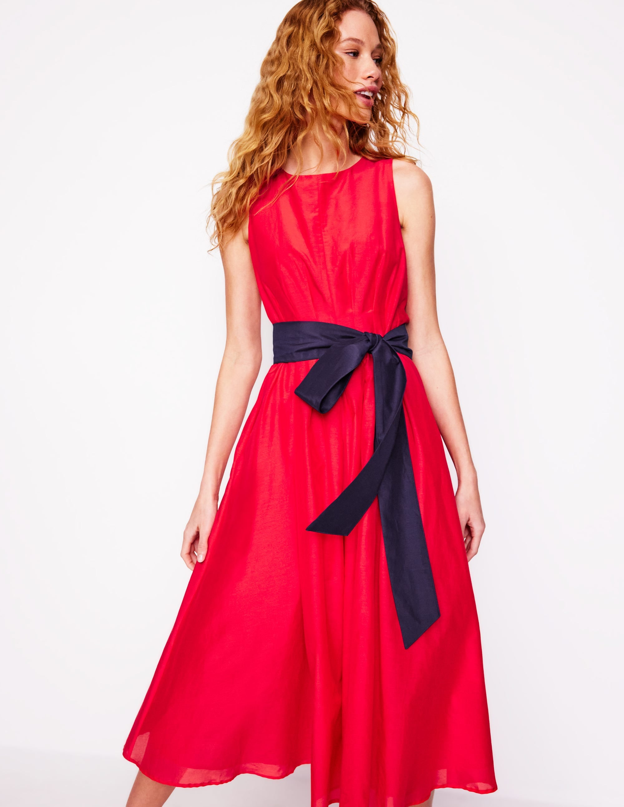 A woman wearing a sleeveless, pleated, poppy red dress with a navy fabric belt tied in a bow at the waist, mid-calf length, and side seam pockets.