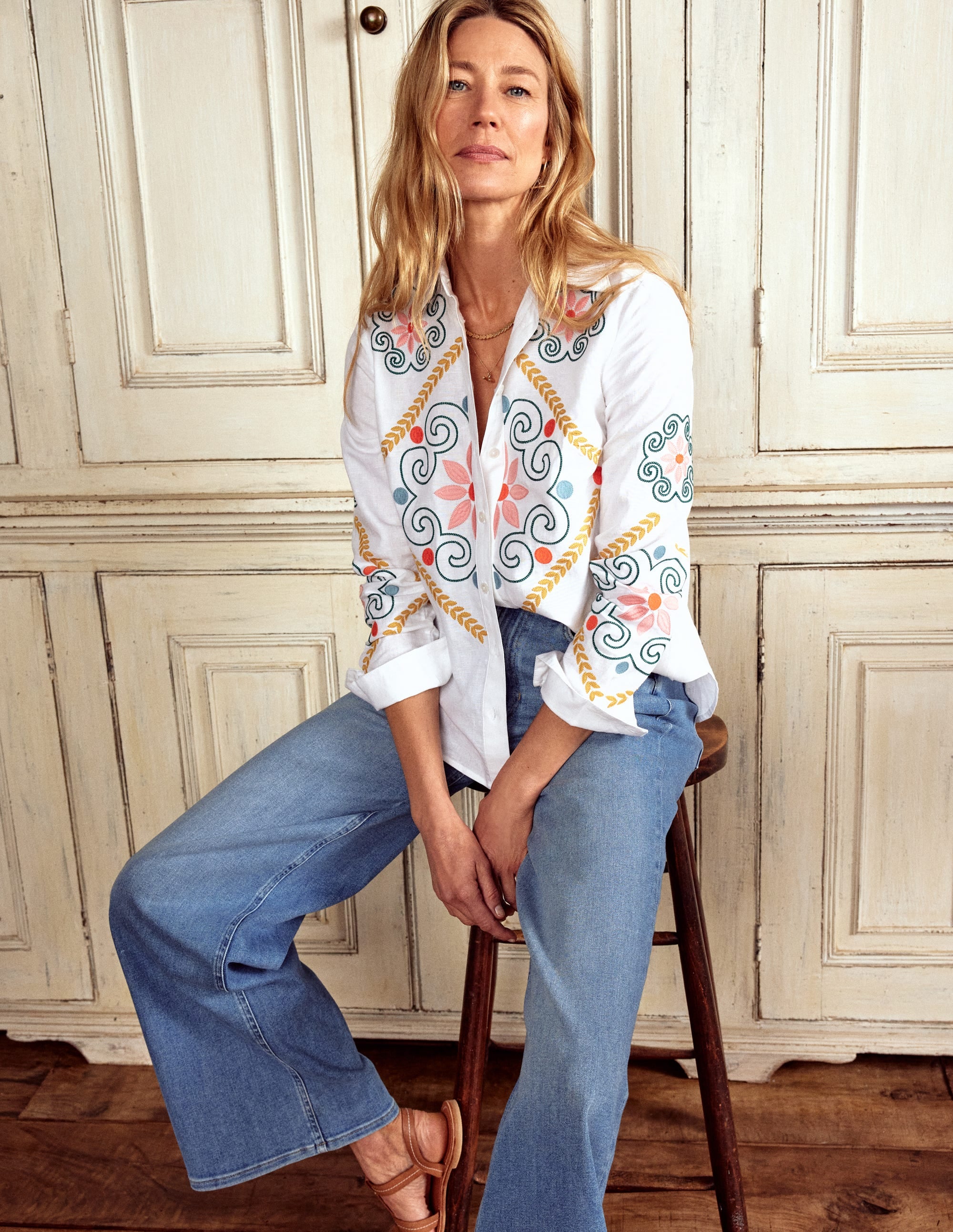 A woman wearing a white embroidered linen shirt with floral and swirl patterns, paired with blue jeans, sitting on a wooden stool.
