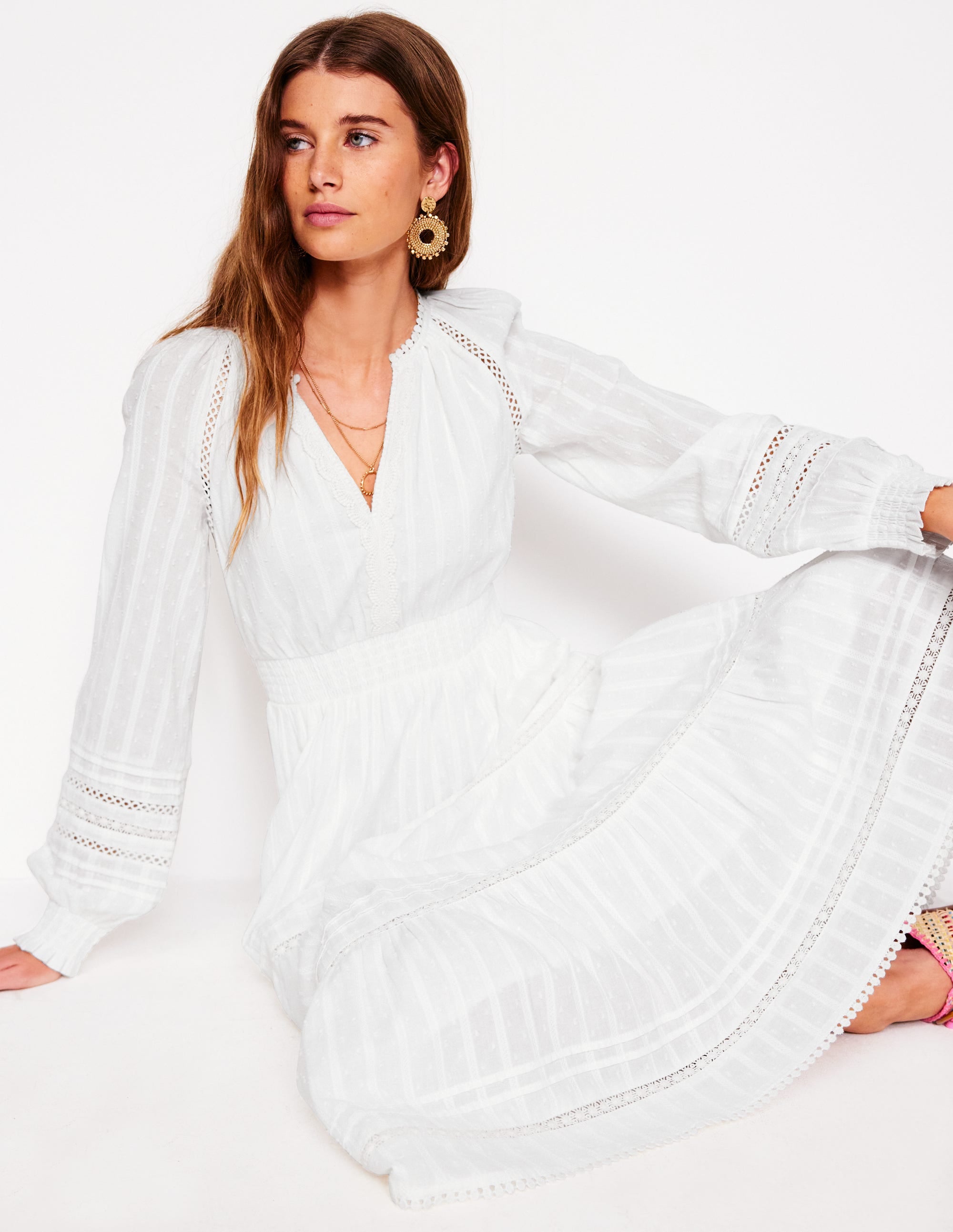 A woman wearing a white, tiered, long-sleeve cotton dress with a V-neck and side pockets.