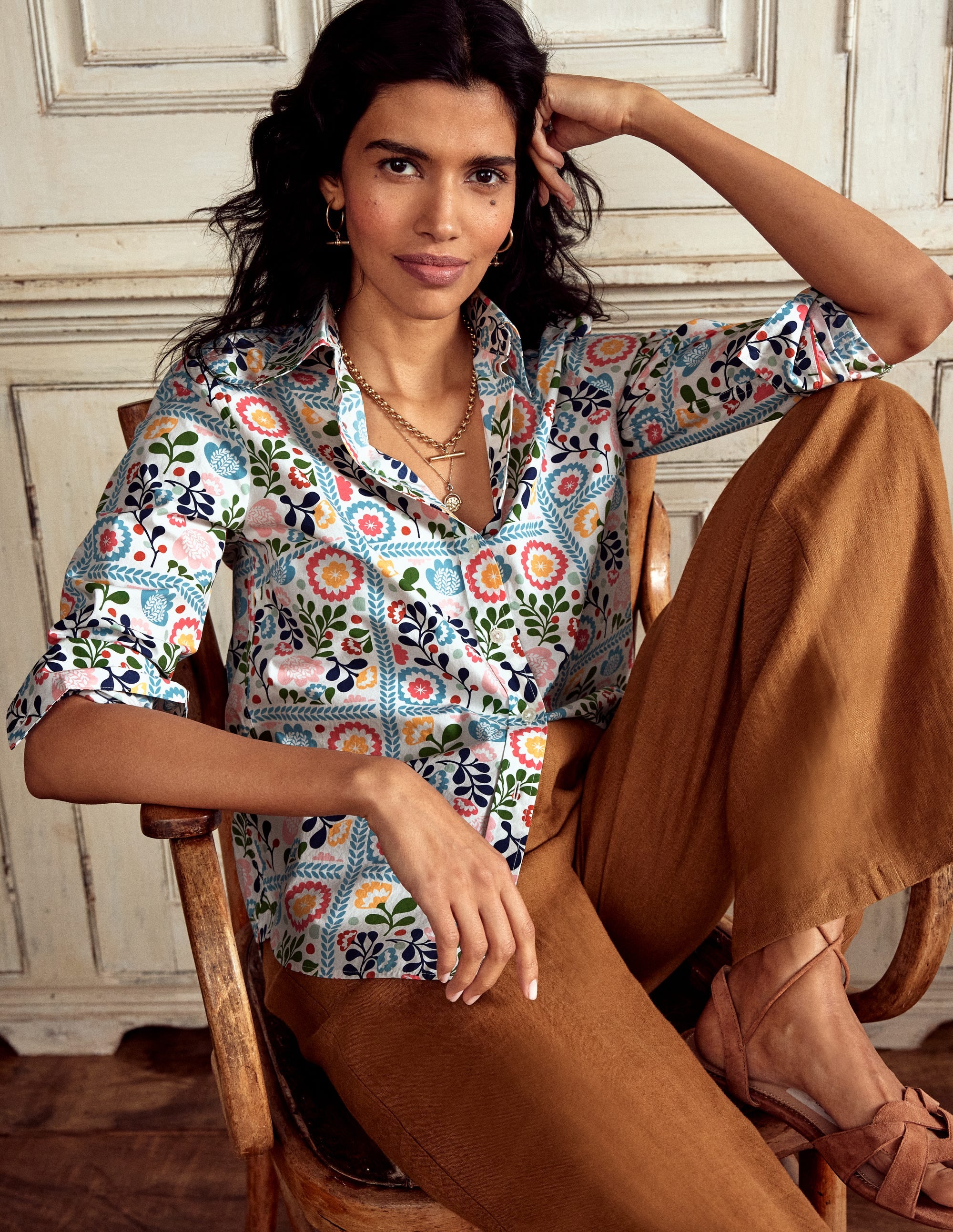 A woman wearing a multicolour vine tile patterned cotton shirt with a collar and long sleeves, paired with brown pants, sitting on a wooden chair.