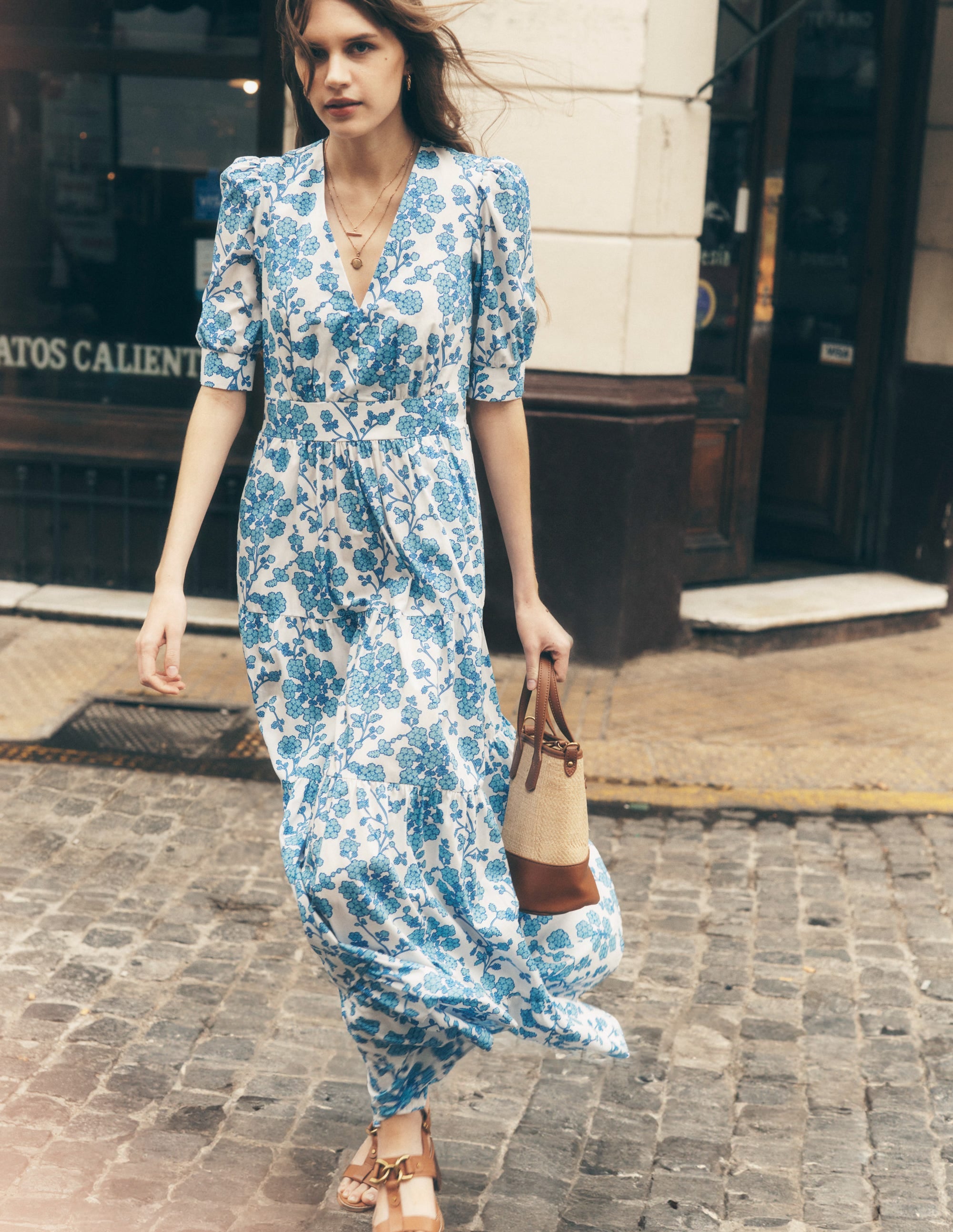 A woman wearing a white and blue floral maxi dress with puff sleeves, V-neckline, tiered skirt, and side pockets, walking on a cobblestone street.
