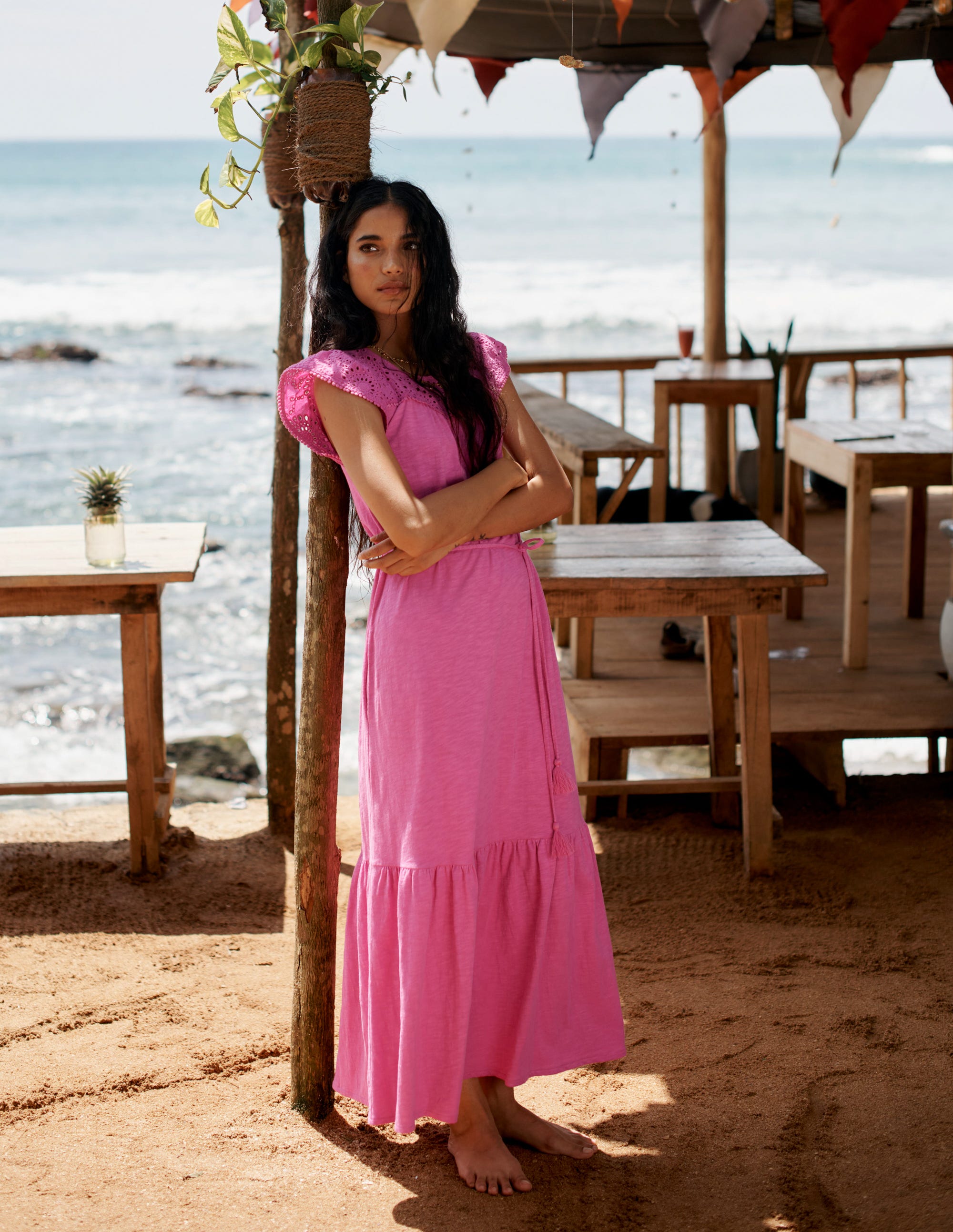 A woman wearing a pink midi dress with flutter sleeves, a tassel belt, and a keyhole back fastening, standing on a sandy beach with wooden tables and the ocean in the background.