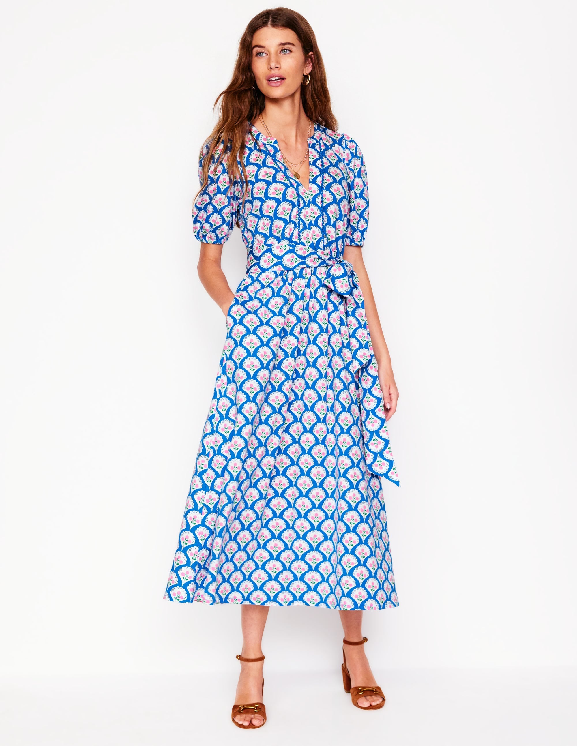 A woman wearing a bright blue midi dress with puff sleeves, scallop trim, and a self-fabric belt, standing against a white background.