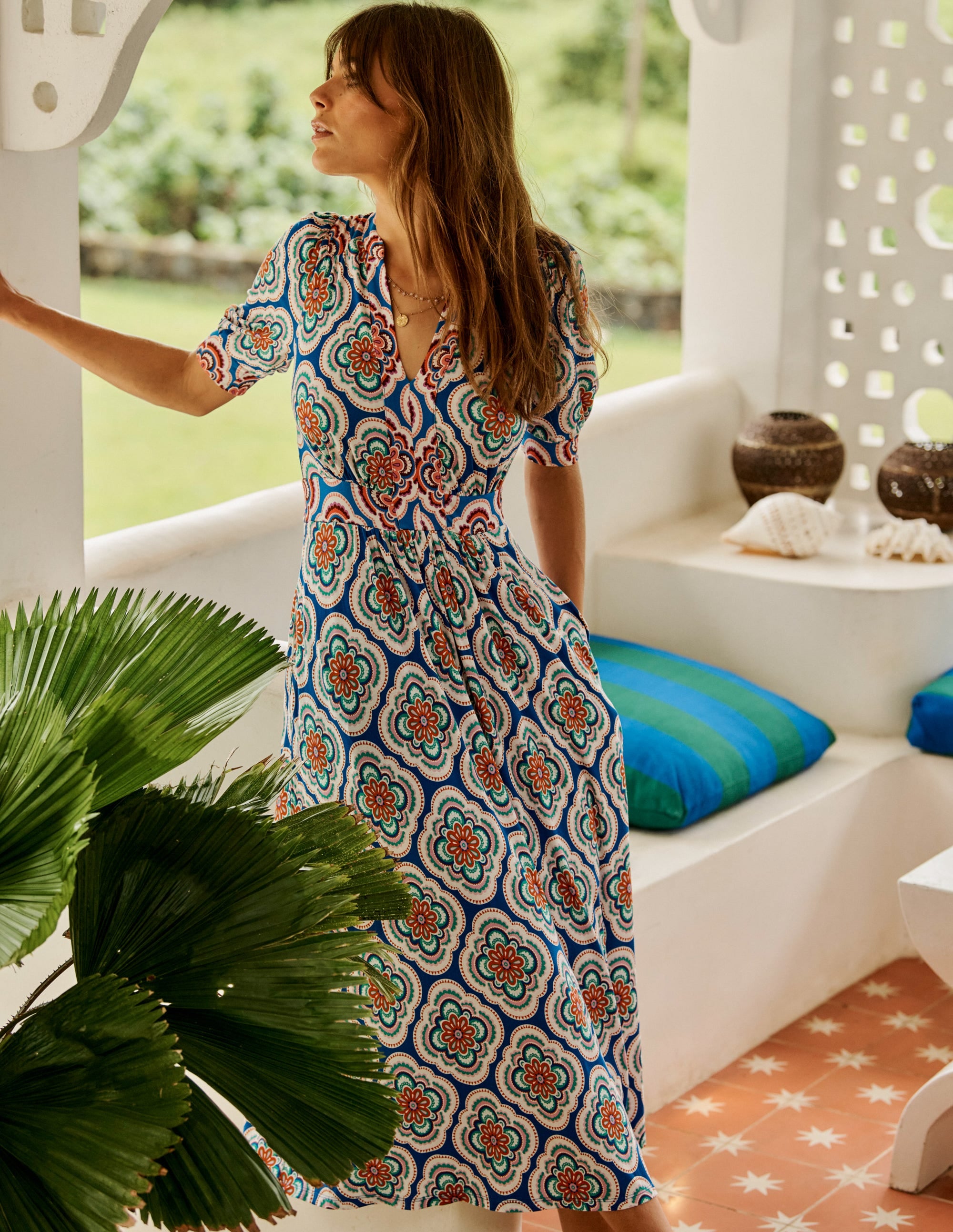 A woman wearing a bright blue midi dress with a colorful floral pattern, puff sleeves, and a flowing skirt, standing indoors with a garden view.
