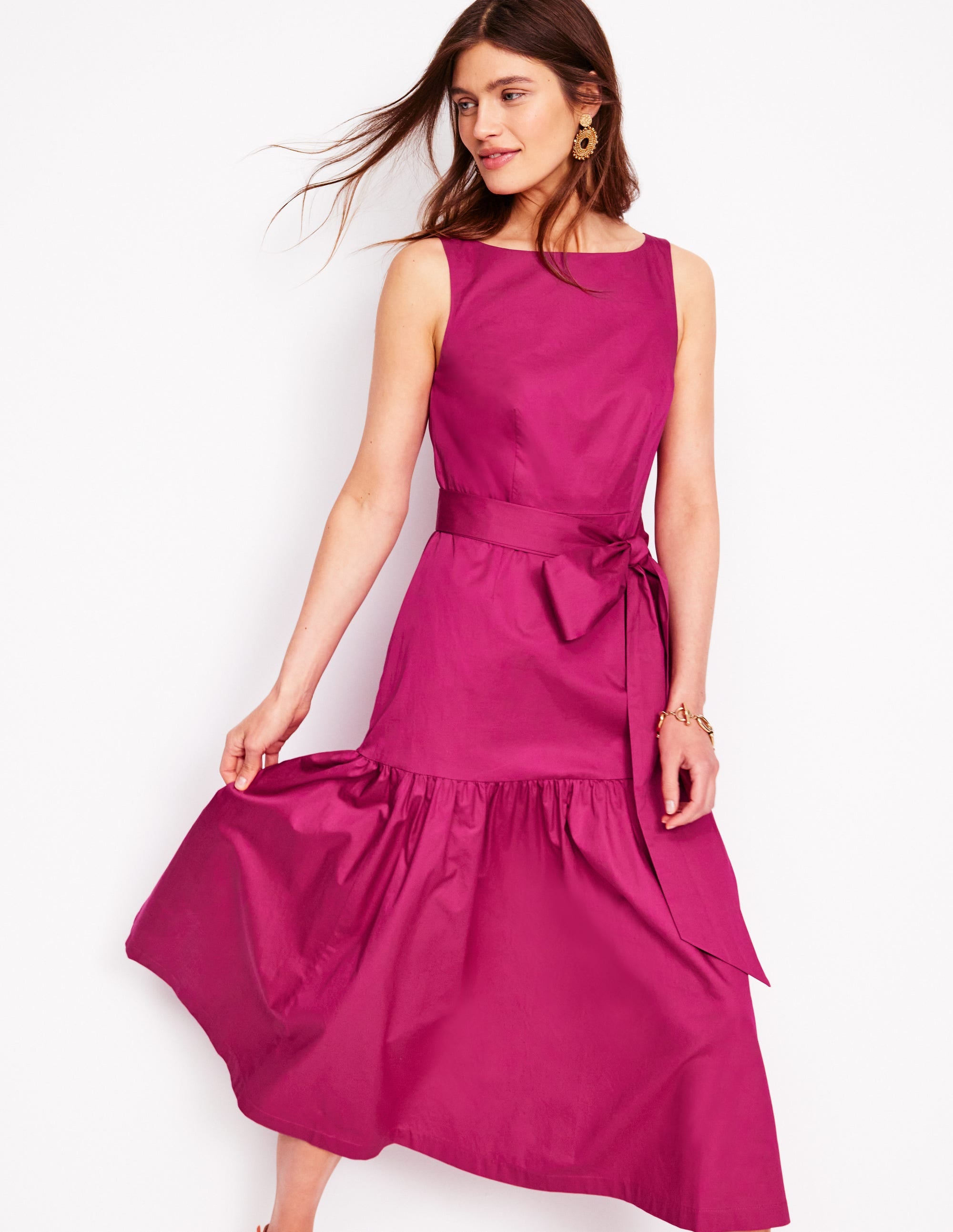 A woman wearing a sleeveless, pink fit and flare maxi dress with a waist tie and side pockets.