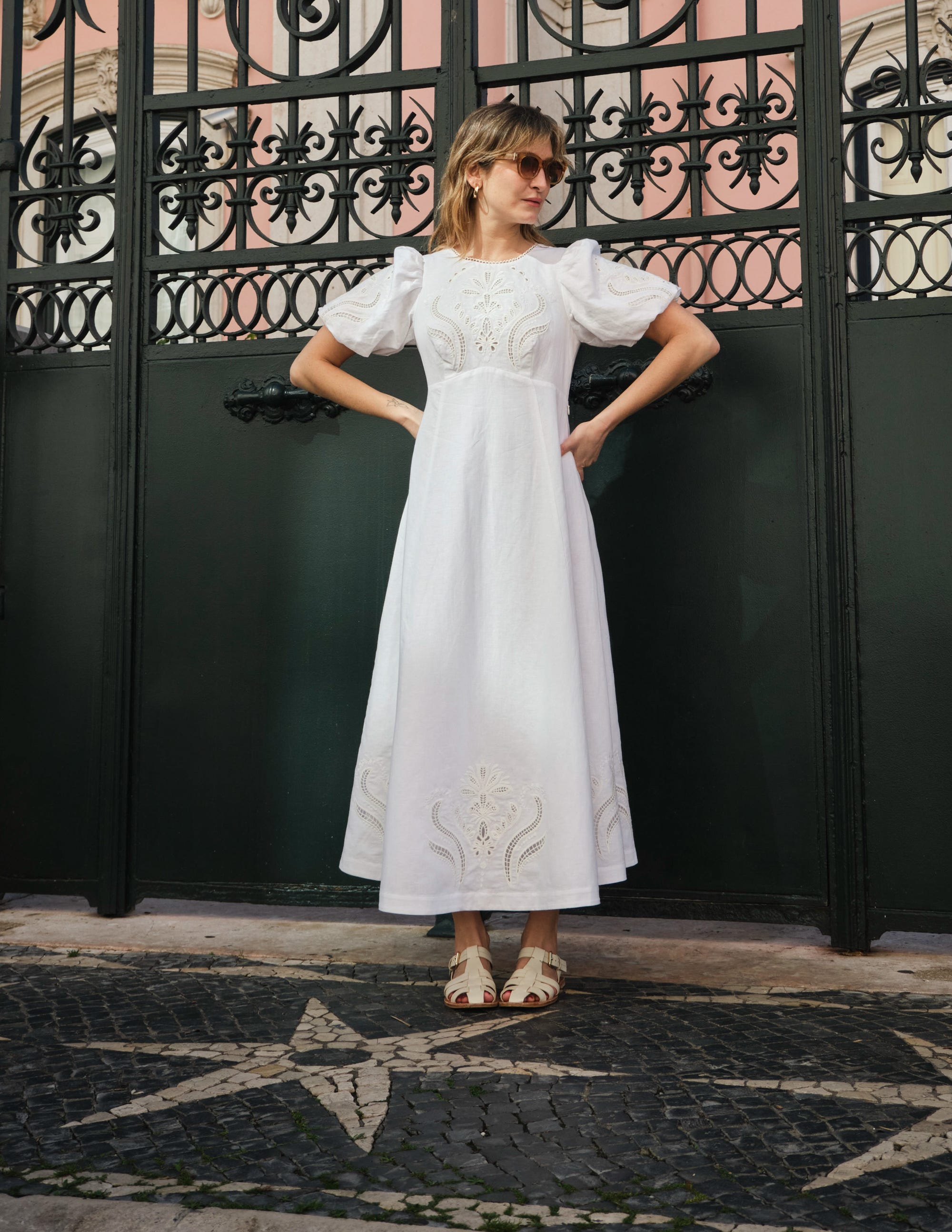 A woman wearing a white empire broderie tea dress with puff sleeves, standing in front of a decorative black gate.