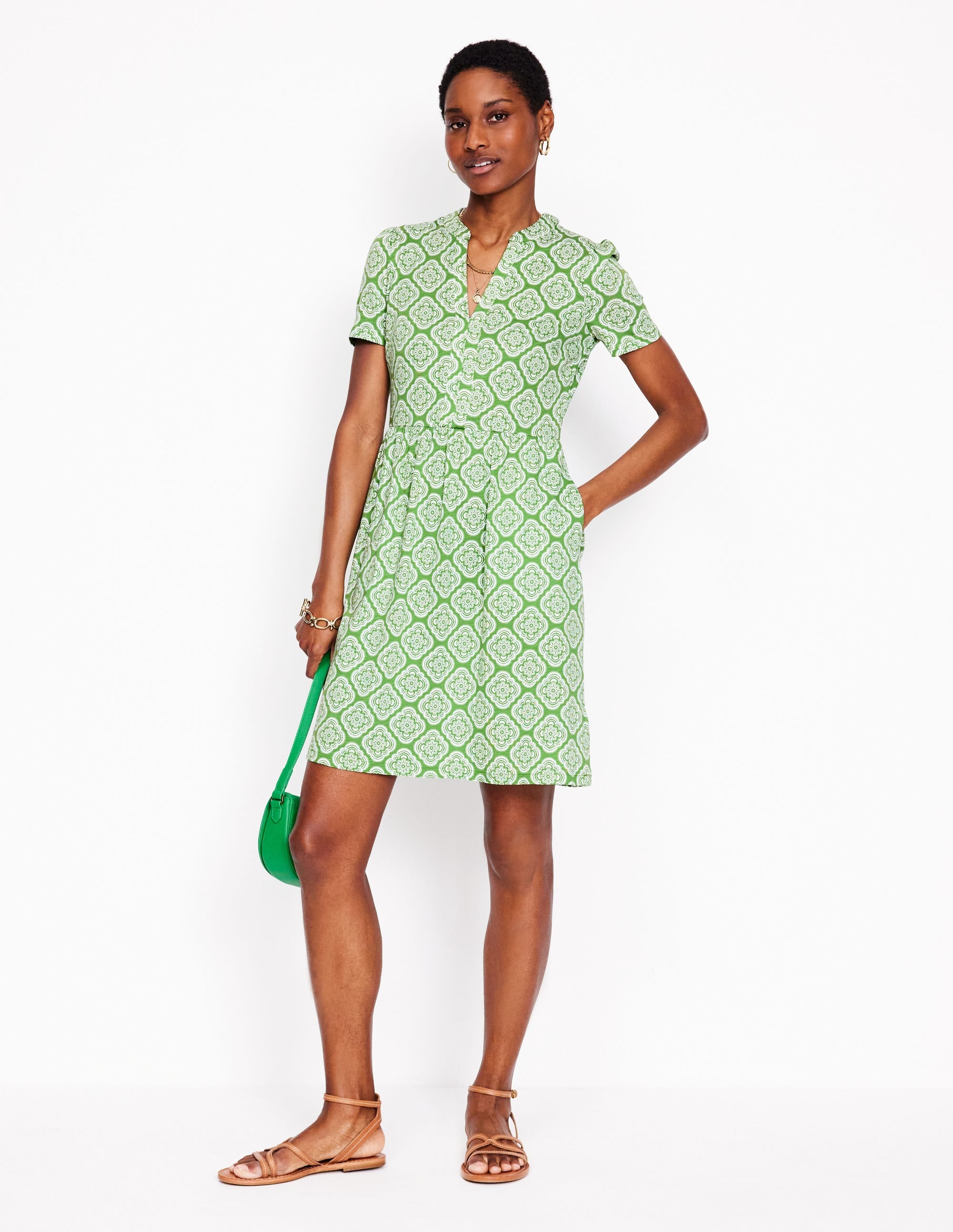 A woman wearing a Paradise Green floral patterned short sleeve dress with a gathered waist and above-the-knee length, paired with sandals and a green handbag.