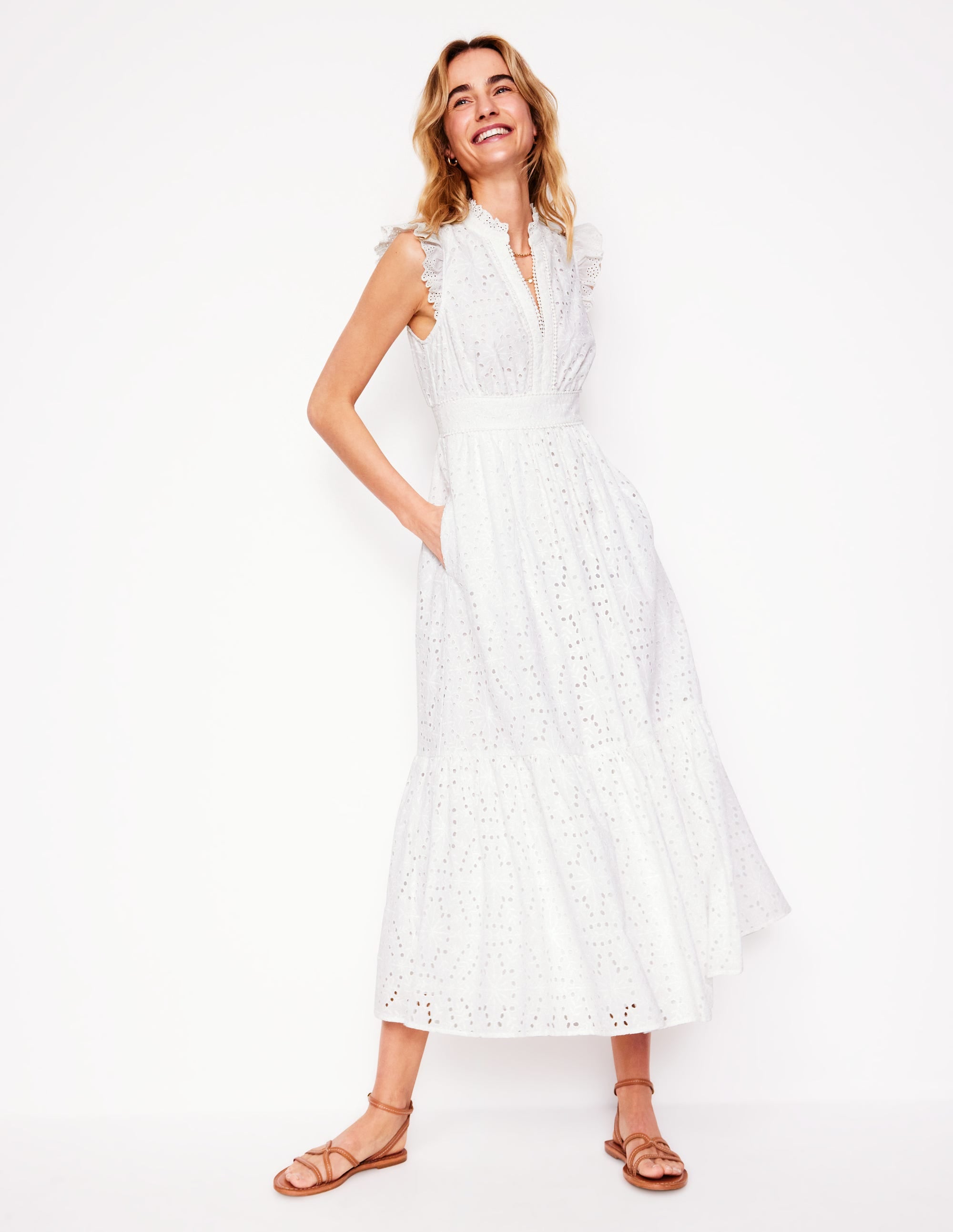 A woman wearing an ivory broderie midi dress with ruffles, pockets, and a smocked back waist, standing against a plain background.