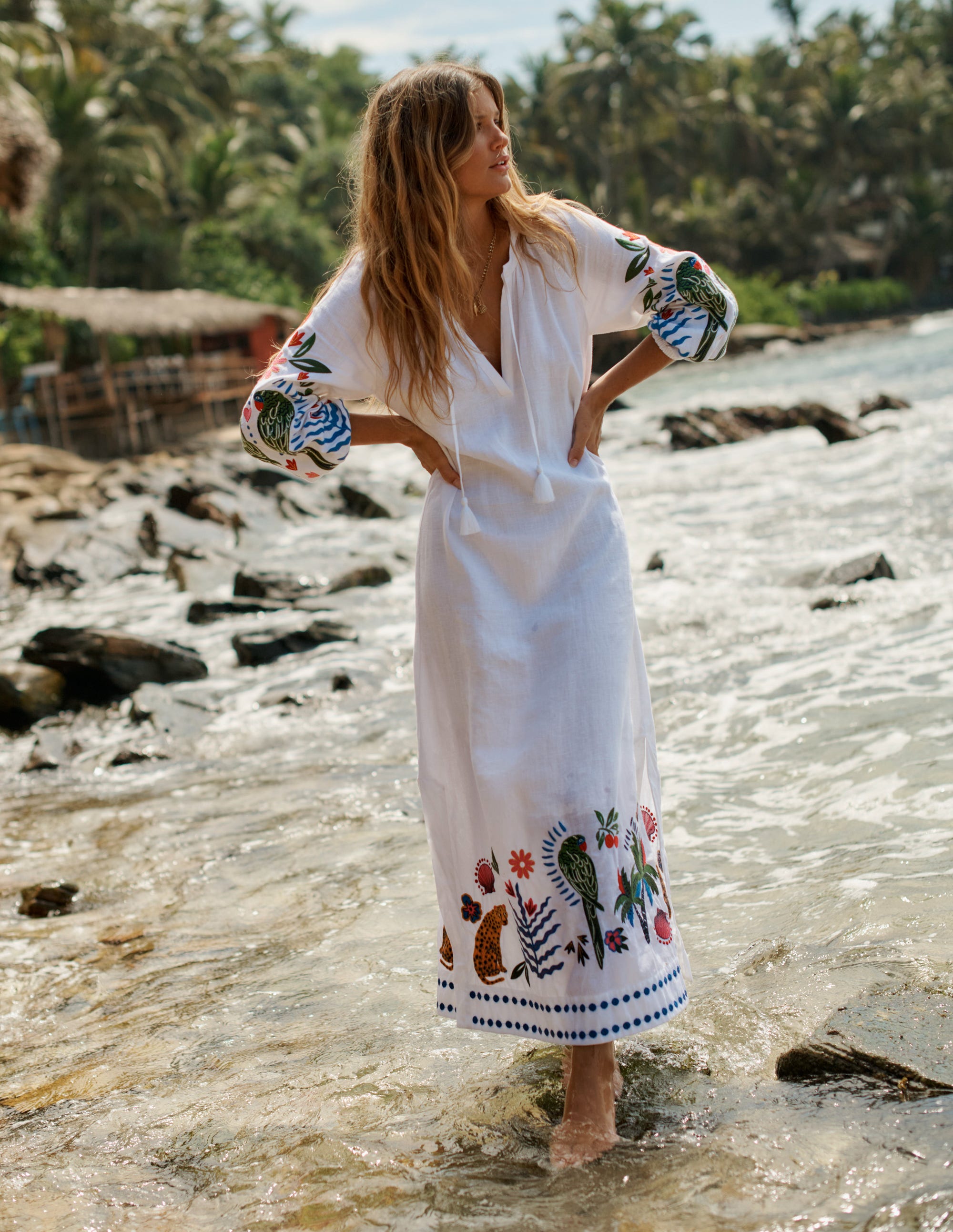 A woman wearing a white linen dress with embroidered and tassel details, standing on a beach with water and rocks.