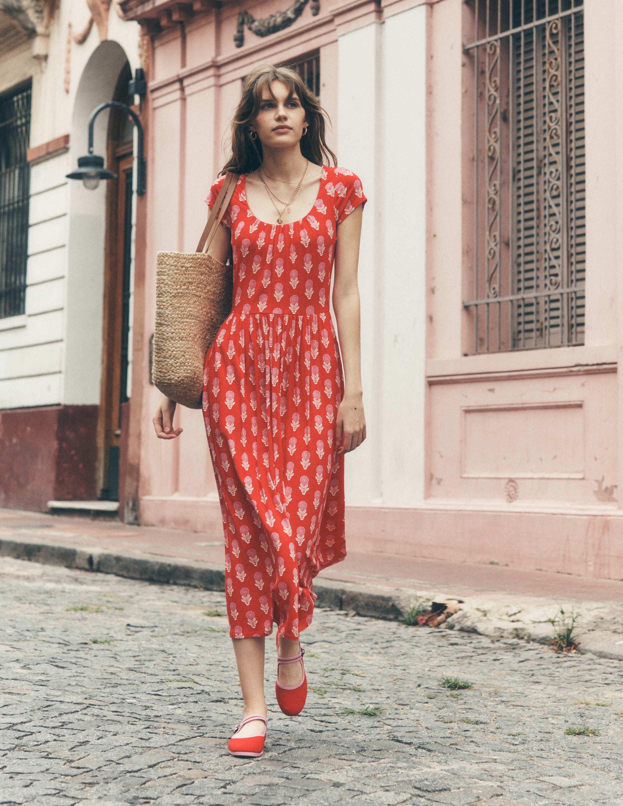 A woman wearing a red dress with a floral print, short sleeves, and a scoop neckline, carrying a woven tote bag, walking on a cobblestone street.