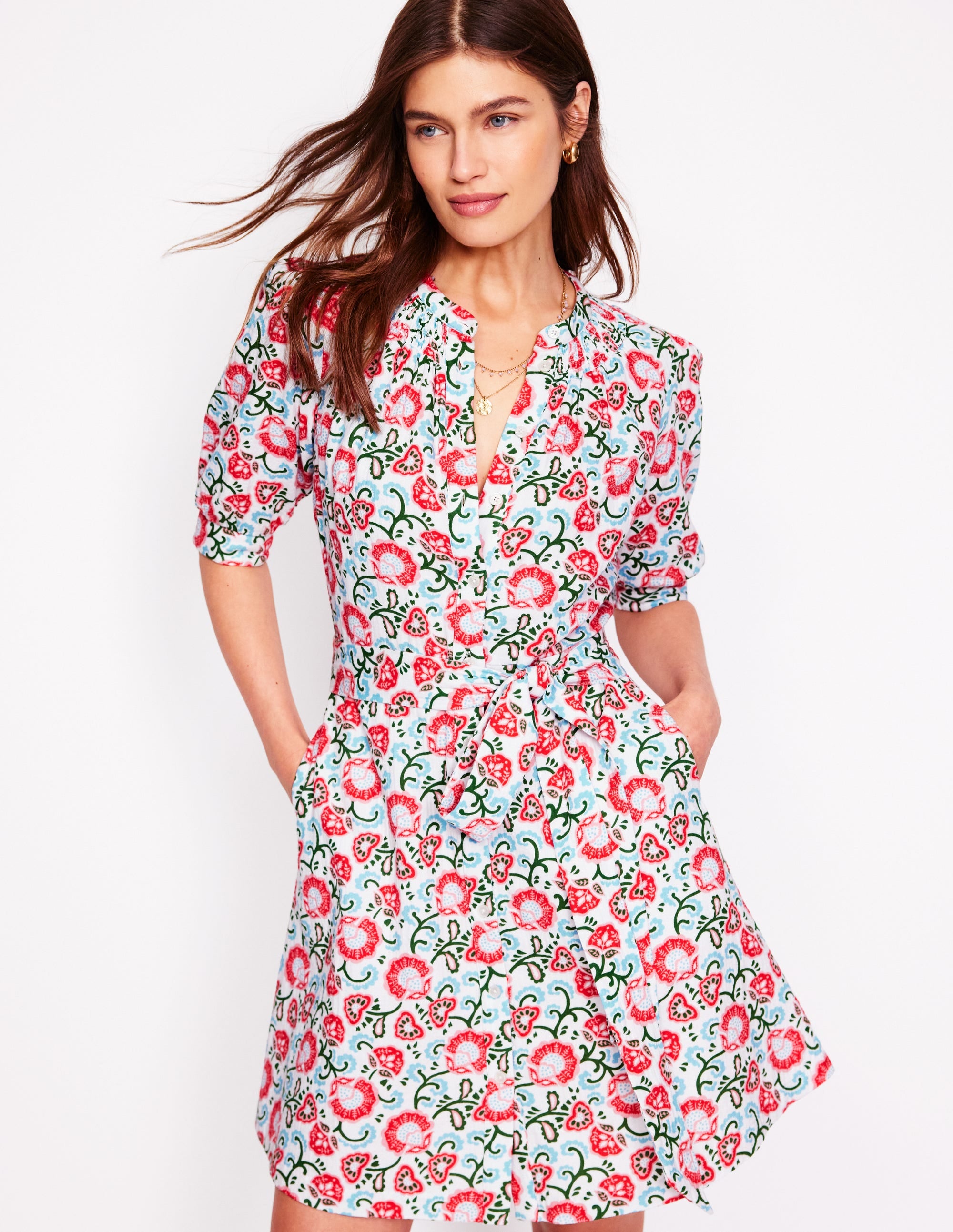A woman wearing an ivory floral dress with gathered sleeves, a smocked neckline, and a self-fabric belt, standing against a white background.