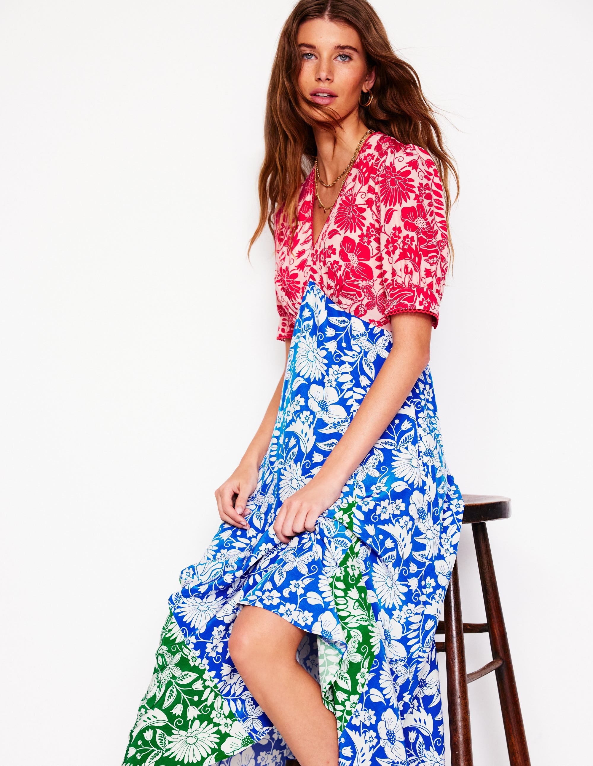 A woman wearing a multicolour floral tea dress with pink, blue, green, and white patterns, featuring a fitted waist, short puff sleeves, and a flowing skirt, sitting on a wooden stool.