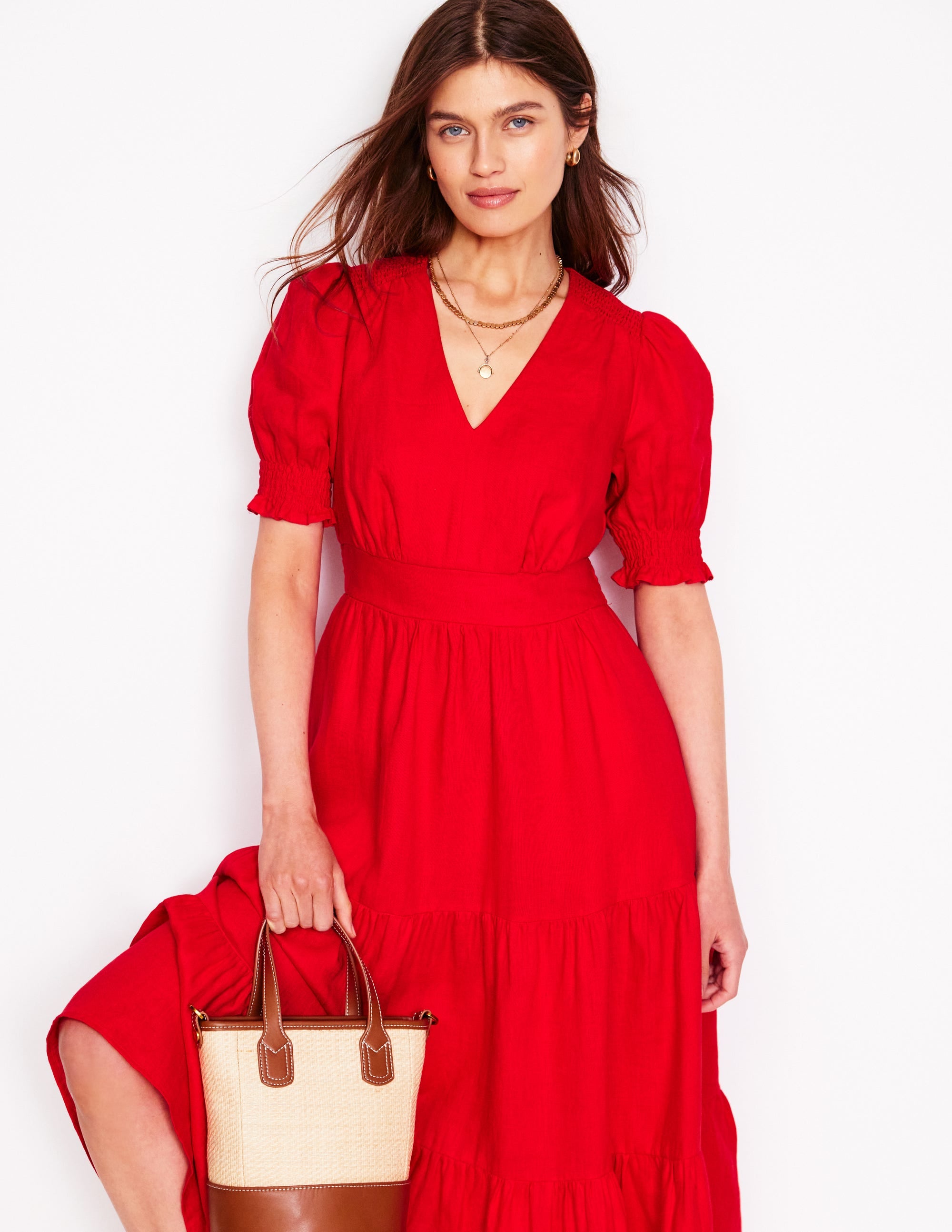 A woman wearing a bright red linen midi dress with puff sleeves and a tiered skirt, holding a beige and brown tote bag.