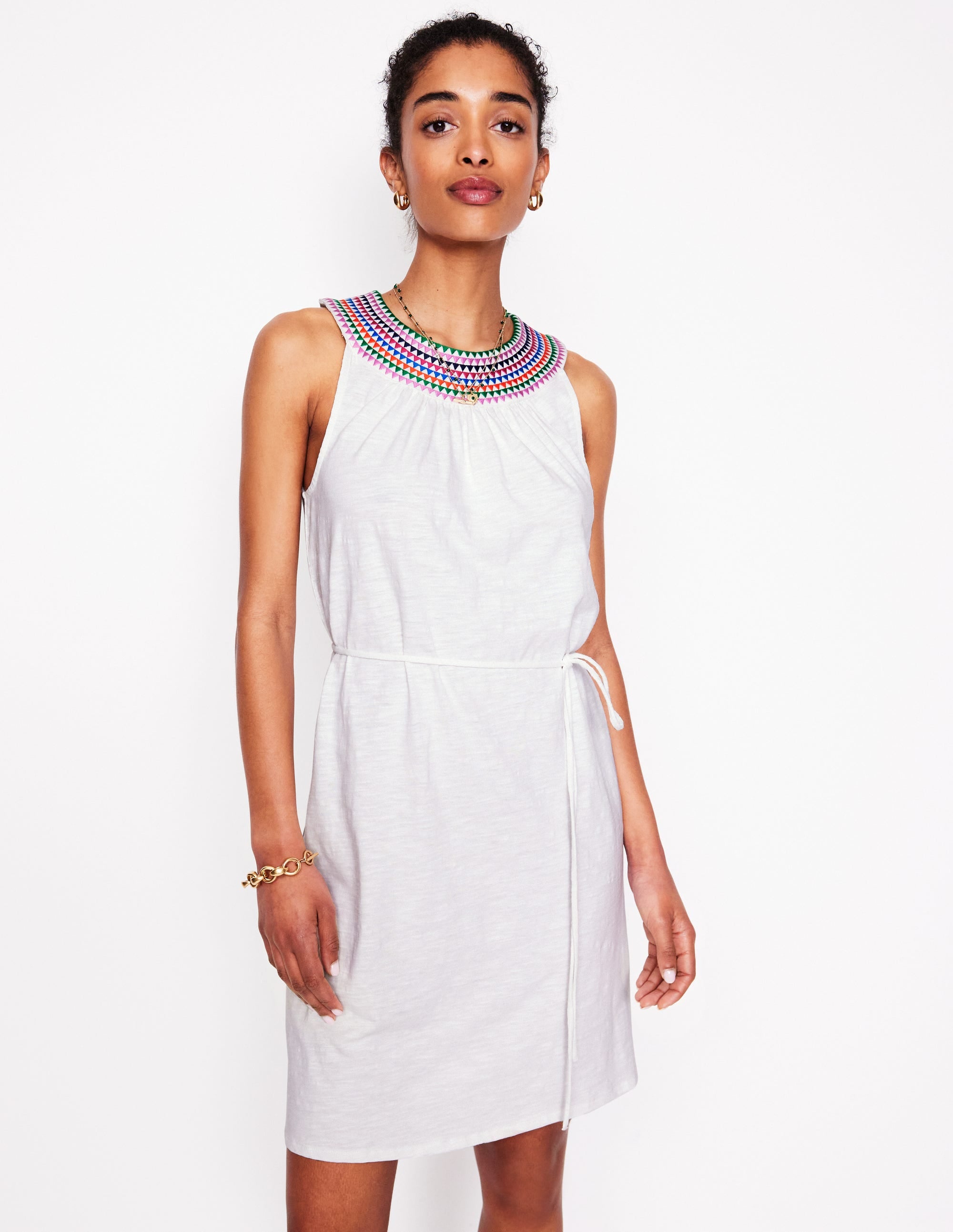 A woman wearing a white embroidered short dress with flutter sleeves and a tie belt, featuring a colorful embroidered neckline.