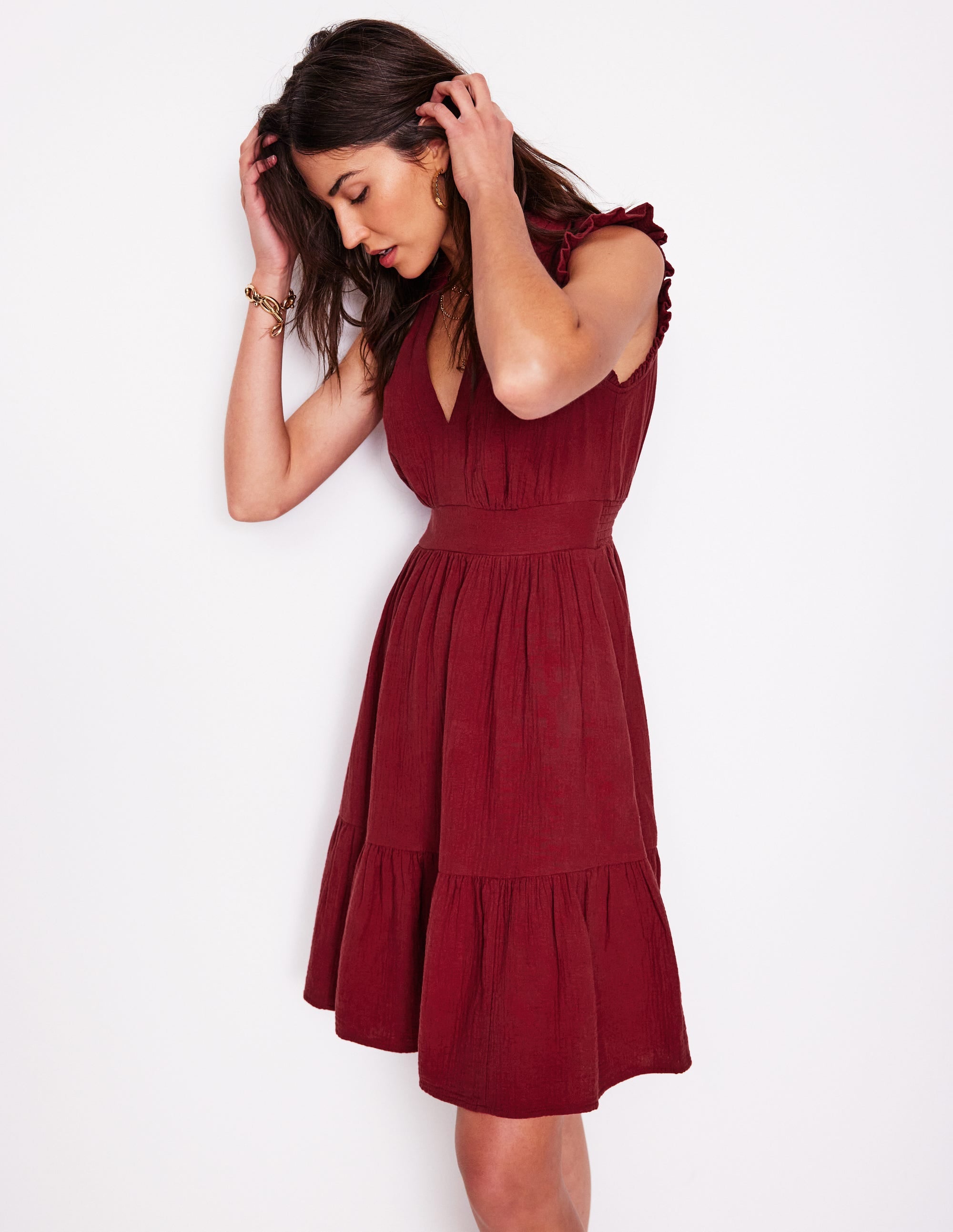 A woman wearing a garnet red short dress with ruffles on the sleeves, a smocked back waist, and a short skirt with pockets.