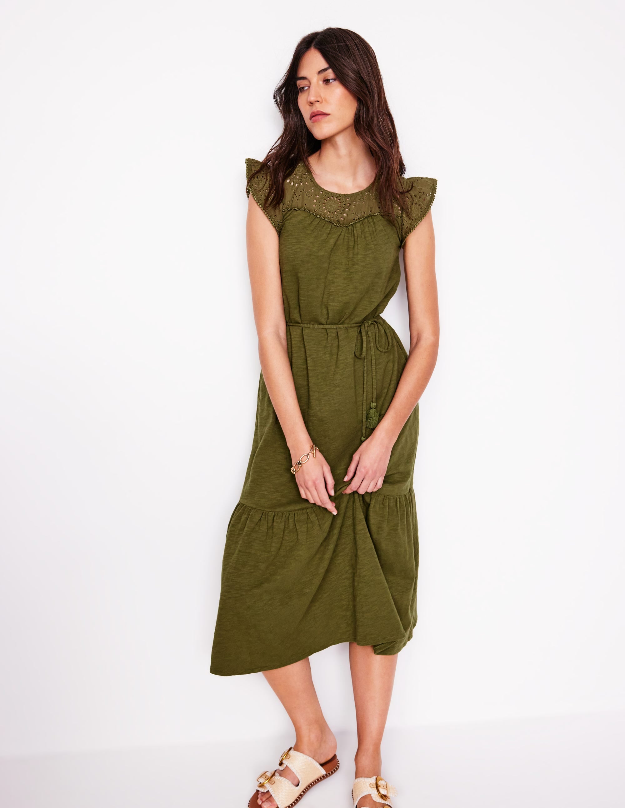 A woman wearing a woodland green midi dress with flutter sleeves, a tassel belt, and woven broderie fabric, standing against a white background.