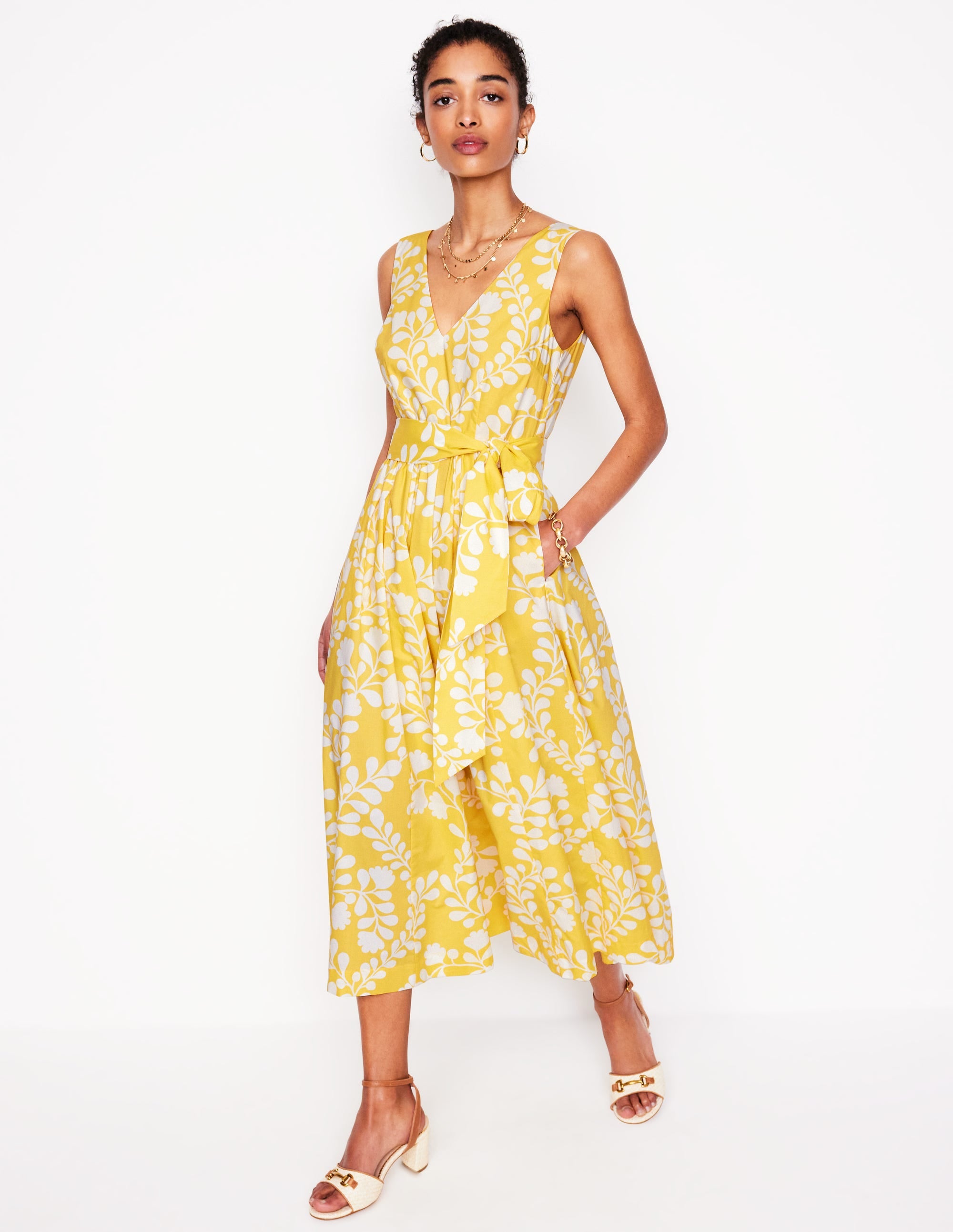A woman wearing a yellow sleeveless V-neck belted flare dress with a white vine floral pattern, midi length, and side pockets.