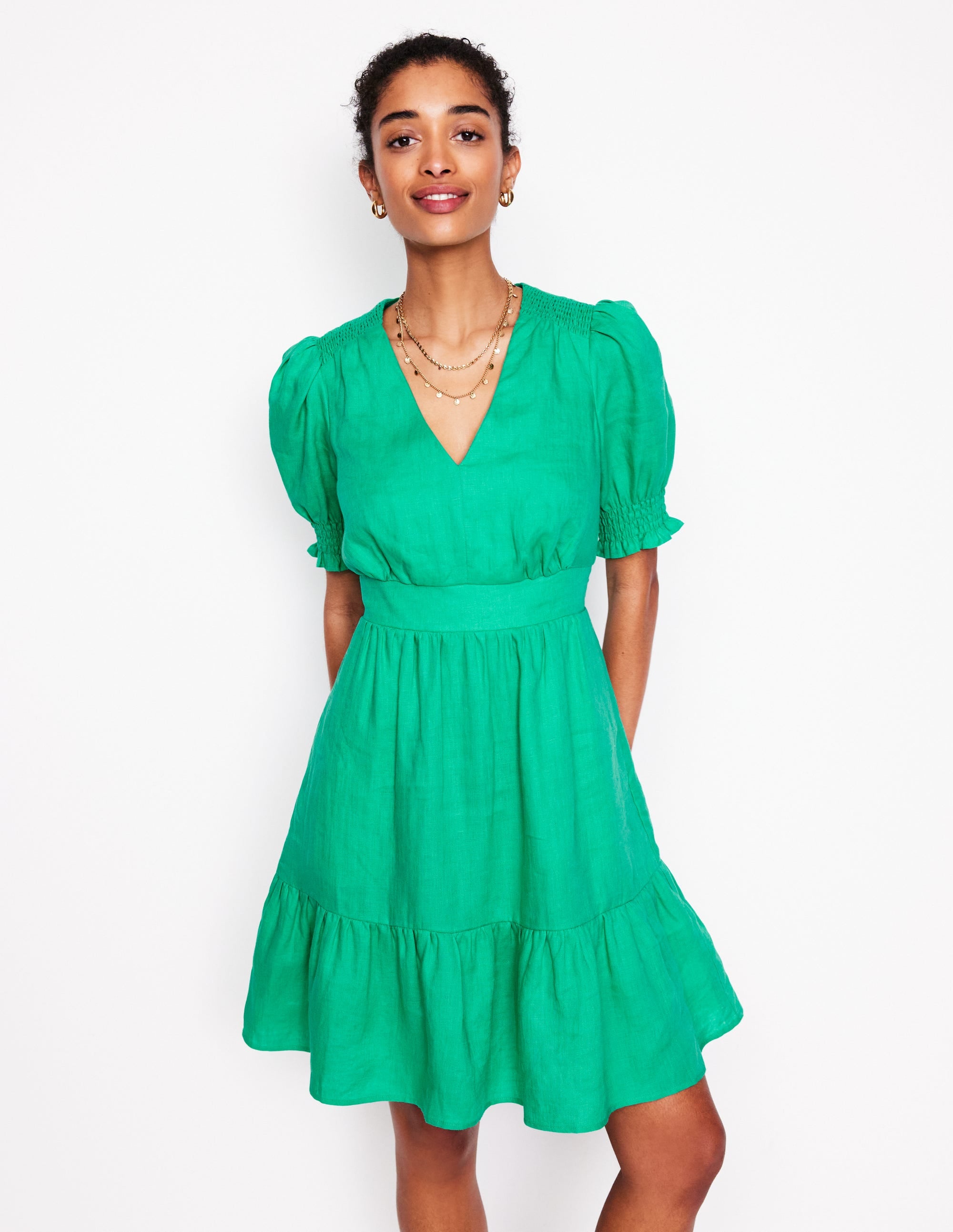 A woman wearing a bright aquamarine blue linen dress with puff sleeves, a V-neckline, and a tiered skirt, standing against a plain white background.