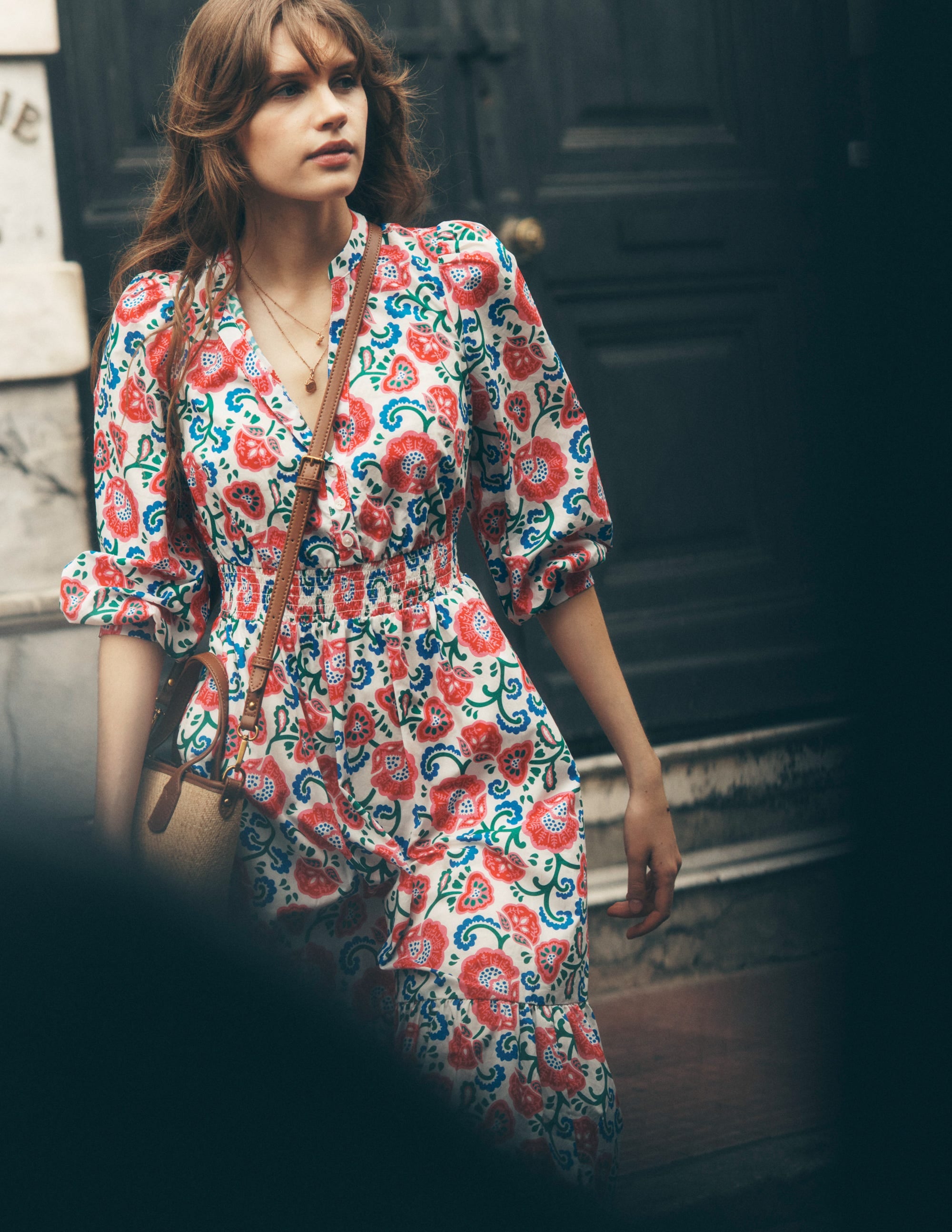 A woman wearing a white cotton midi dress with a vibrant red, blue, and green floral pattern, long puffed sleeves, and a V-neckline.