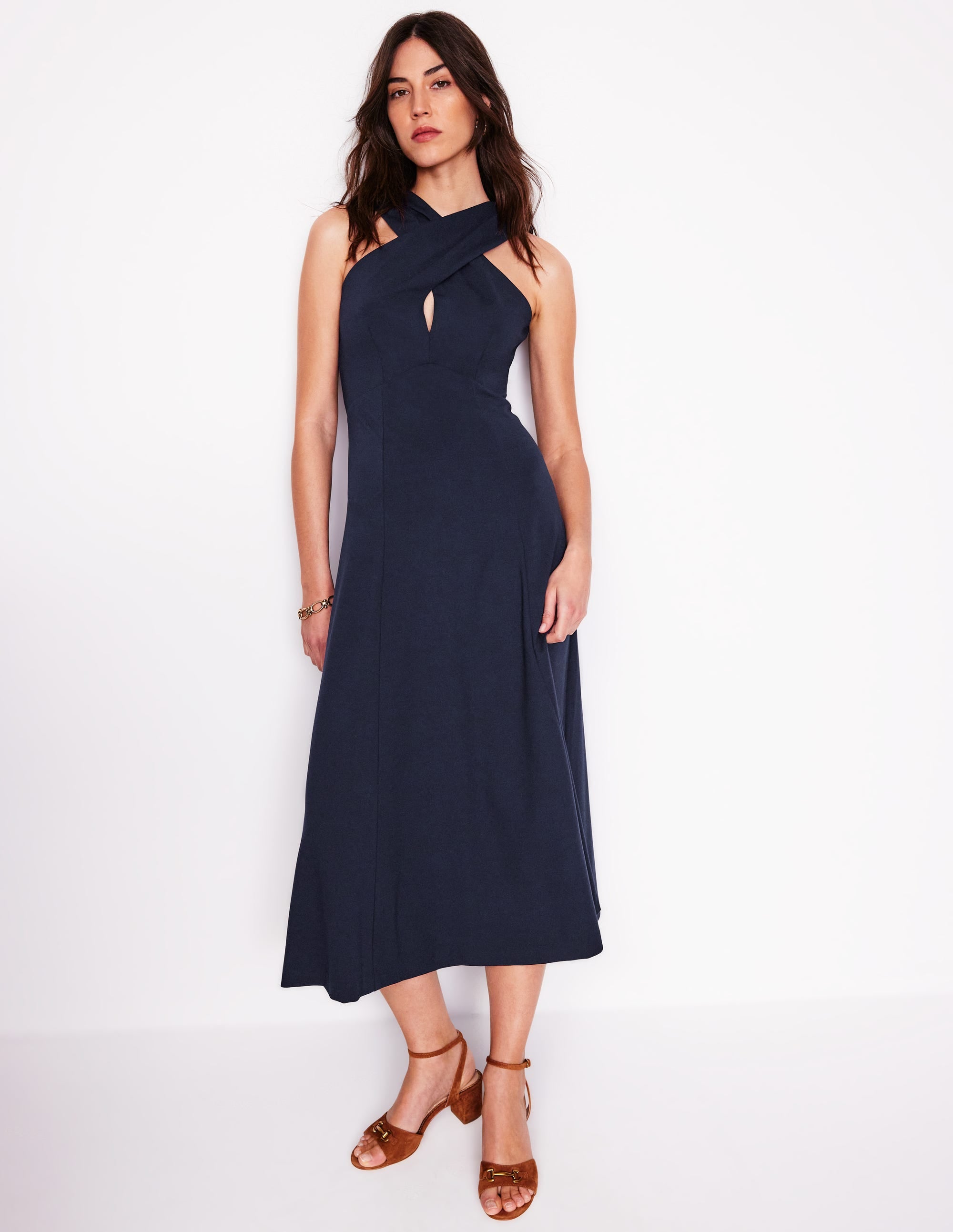 A woman wearing a navy blue, cross-front, fitted maxi dress with a swing silhouette, sleeveless design, and a concealed back zip.