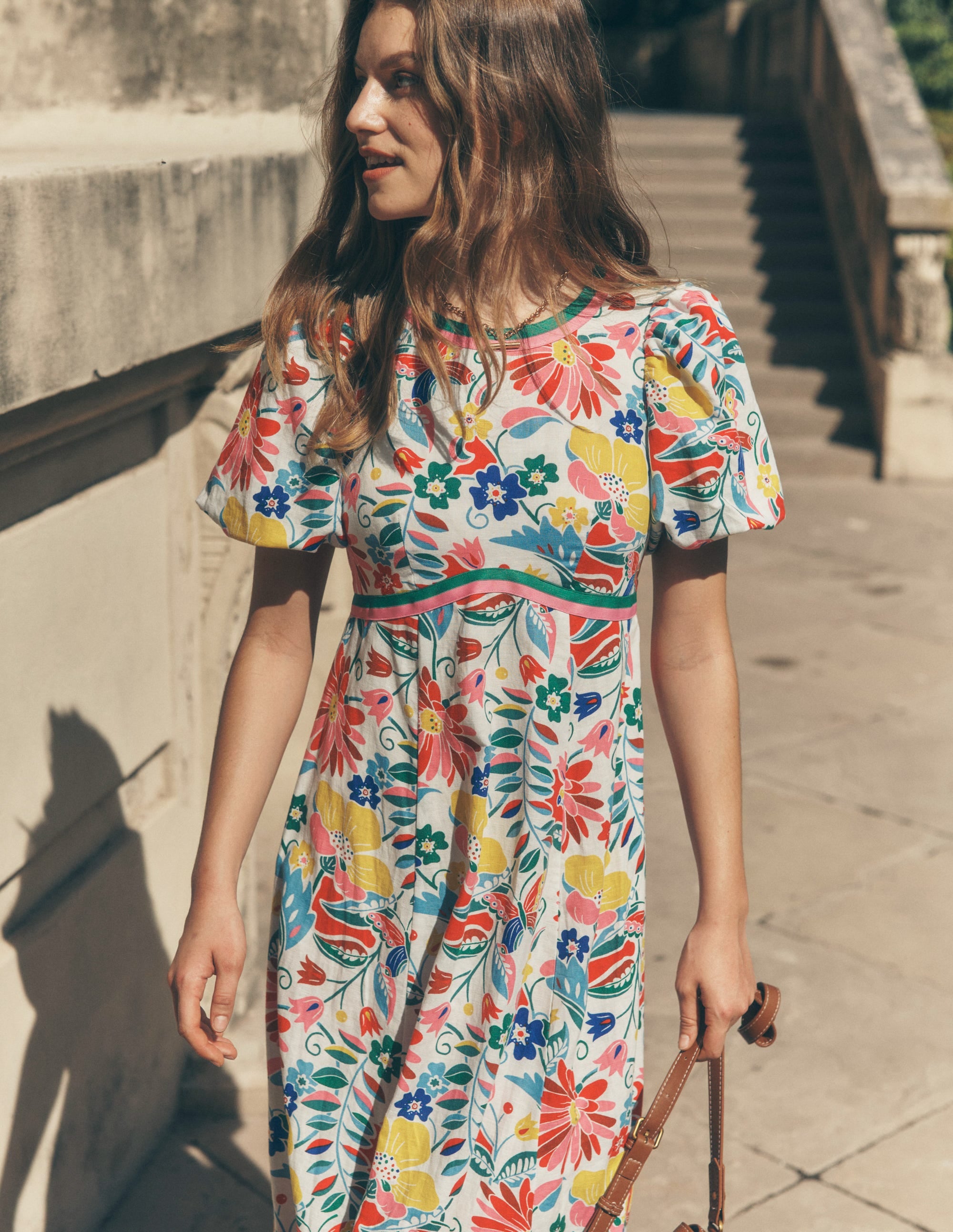 A woman wearing a multi-colored floral empire linen tea dress with puff sleeves, fitted waist, and maxi length, standing outdoors.
