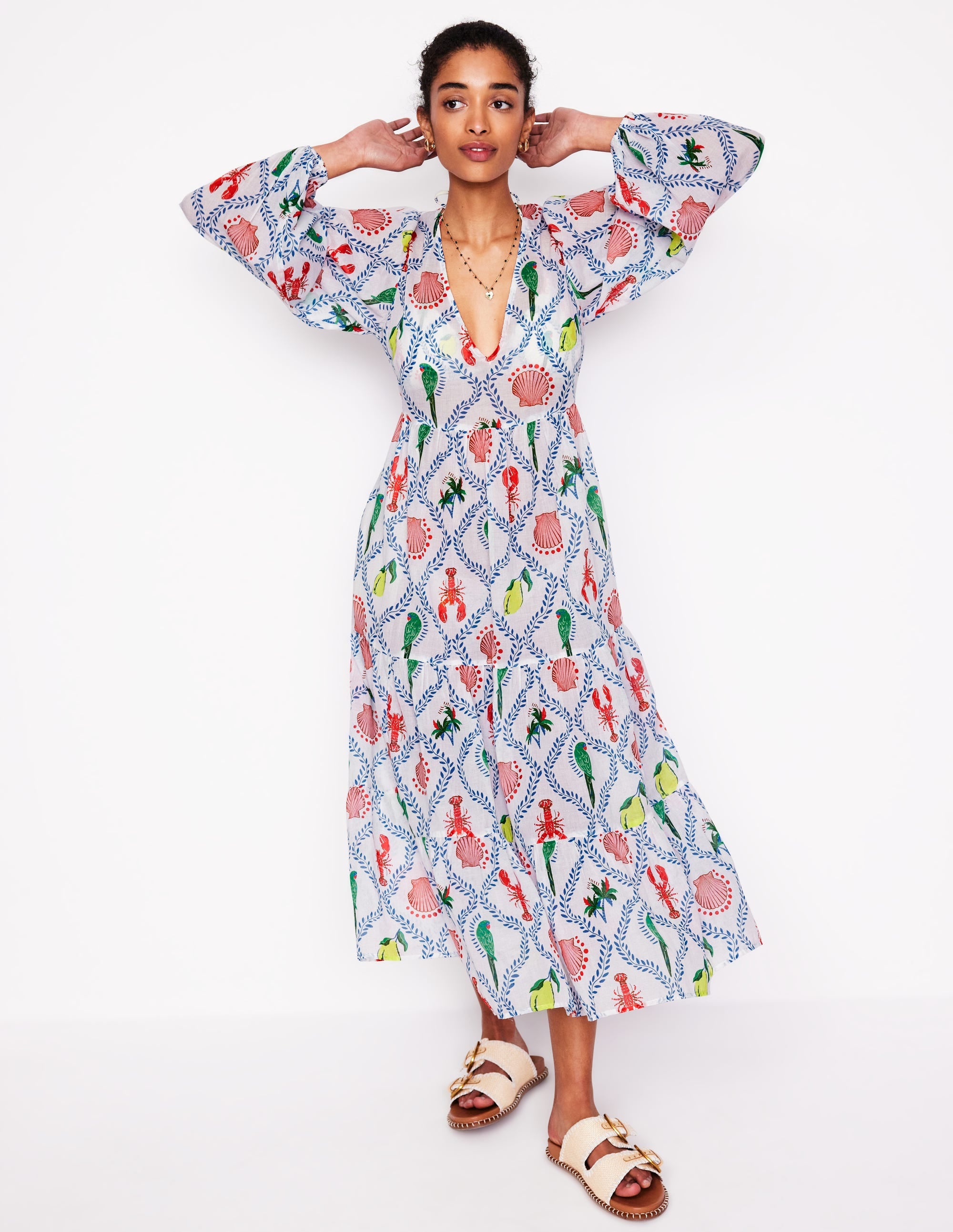 A woman wearing an ivory V-neck tiered cotton kaftan with a colorful tropical print, long sleeves, and side seam pockets.