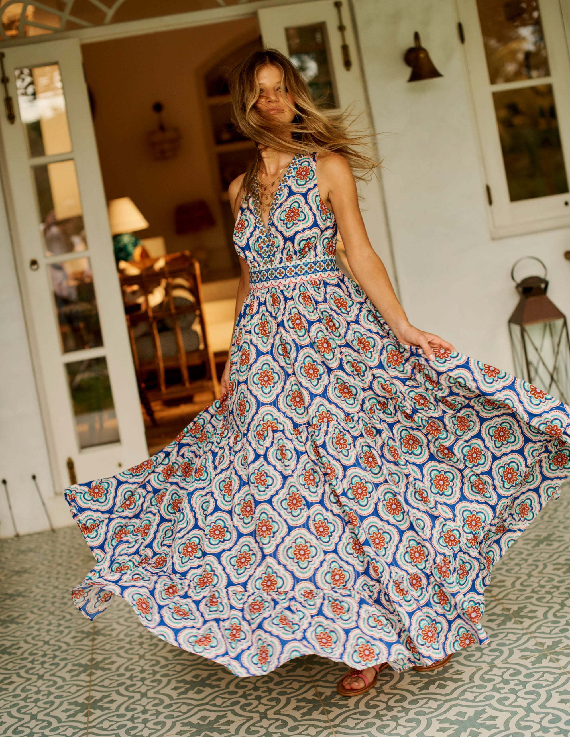 A woman wearing a colorful, patterned maxi dress with a V-neck, scallop trim, and flowing tiers, standing on a patterned floor in a cozy, well-lit room.
