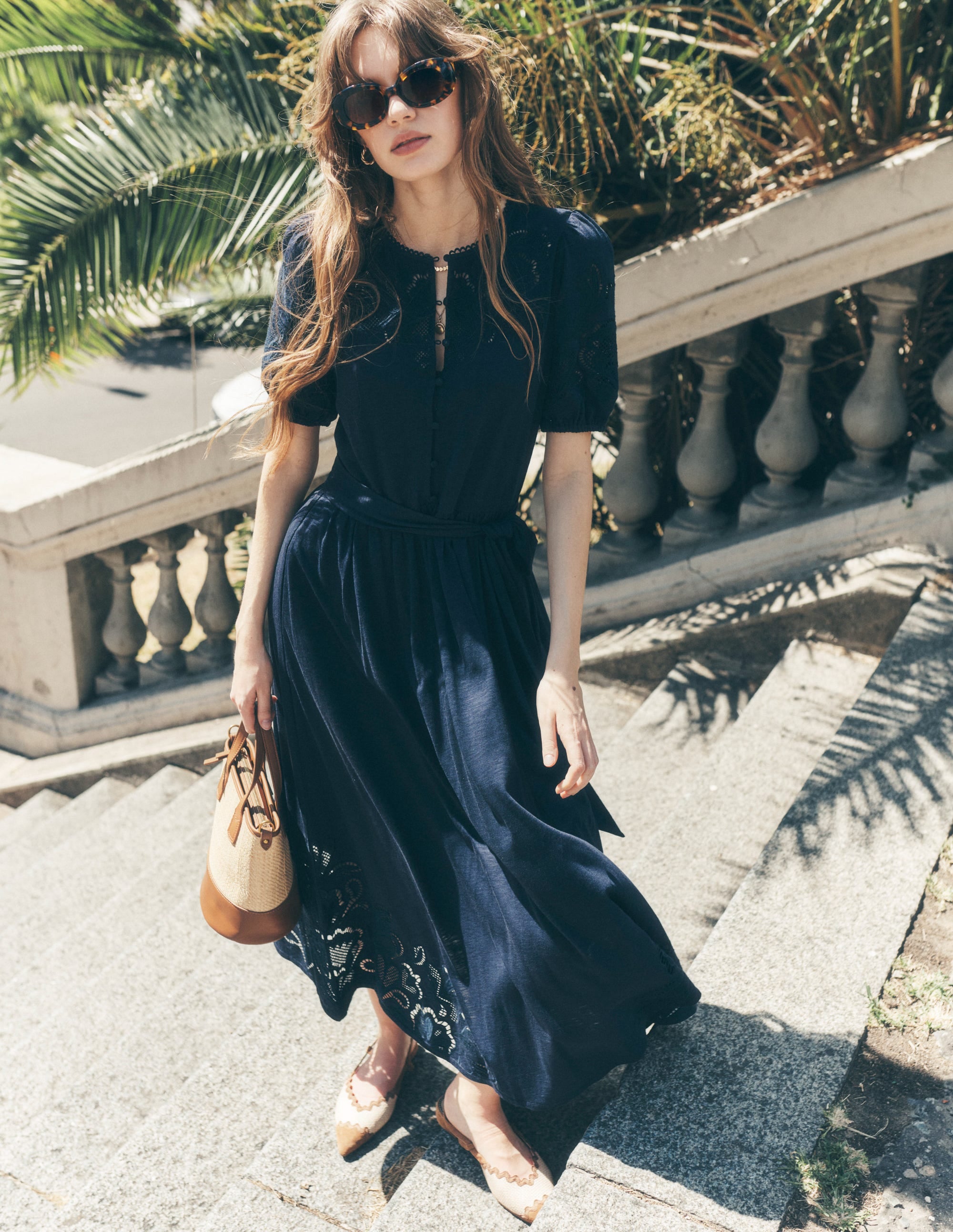 A woman wearing a navy midi dress with cutwork details, puff sleeves, and a fabric belt, standing outdoors near a stone railing with greenery in the background.