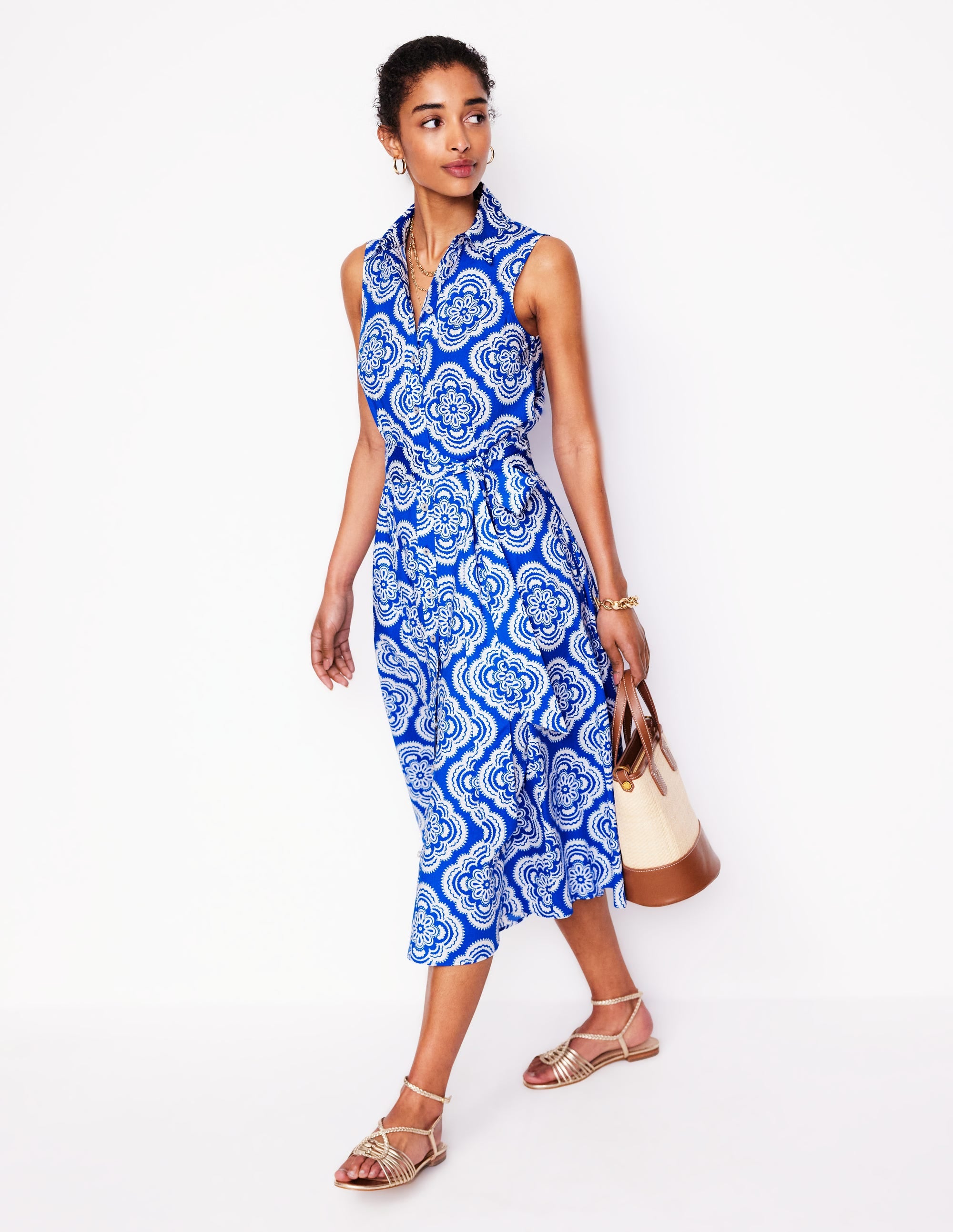 A woman wearing a sleeveless blue and white floral patterned shirt dress with a collar, buttons at the front, and a self-fabric belt, paired with sandals and carrying a beige and brown handbag.