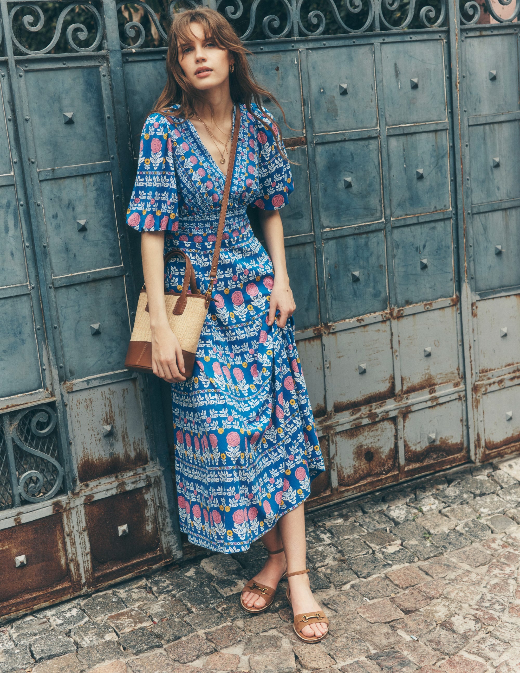 A woman wearing a blue floral midi dress with puff sleeves and a V-neckline, standing against a metal gate.