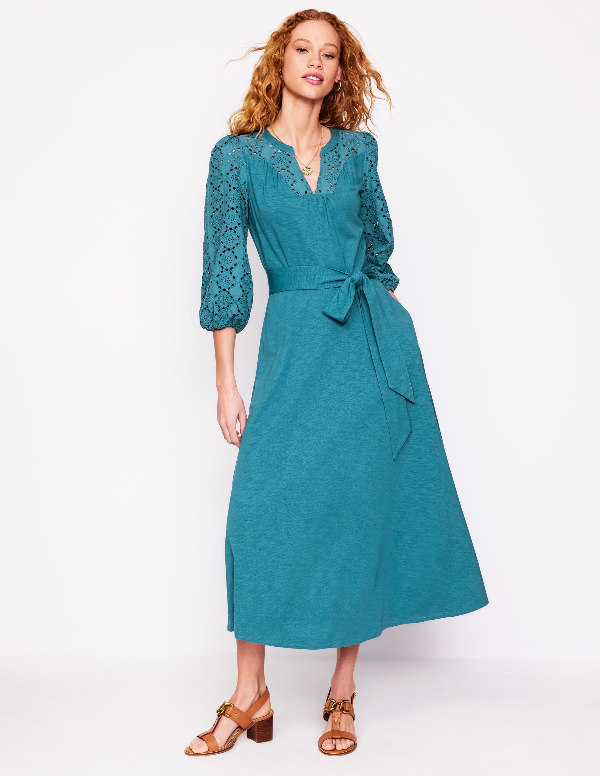 A woman wearing a Waterfall Blue Eden Broderie Jersey Dress with broderie details on the top and sleeves, a waist tie, and a flowing calf-length skirt.