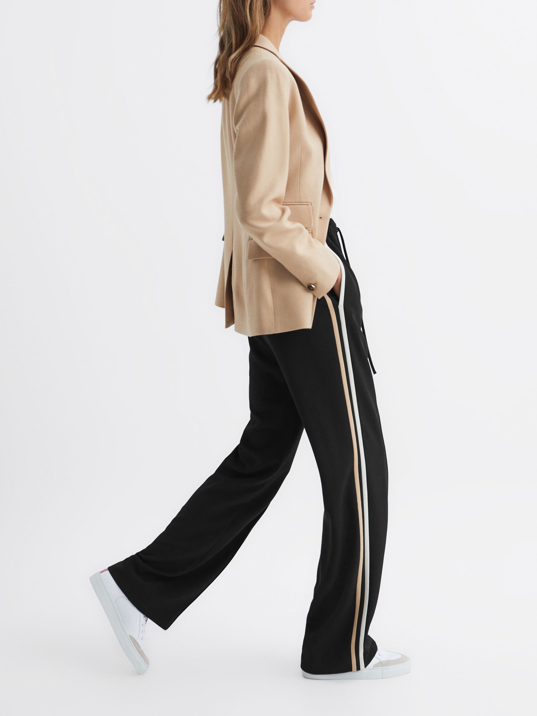 A woman wearing black wide-leg trousers with side stripes, paired with a beige blazer and white sneakers.