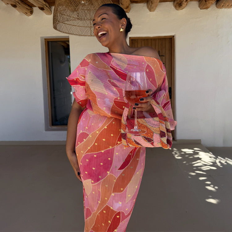 A woman wearing a pink abstract patterned dress with a fitted skirt and a voluminous top, holding a glass of rosé wine, standing in a sunlit room with wooden beams.