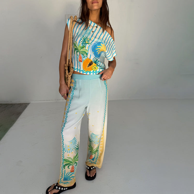 A woman wearing white wide-leg trousers with a colorful Rio print, including pineapples, palm trees, and star motifs, paired with a matching top and sandals.