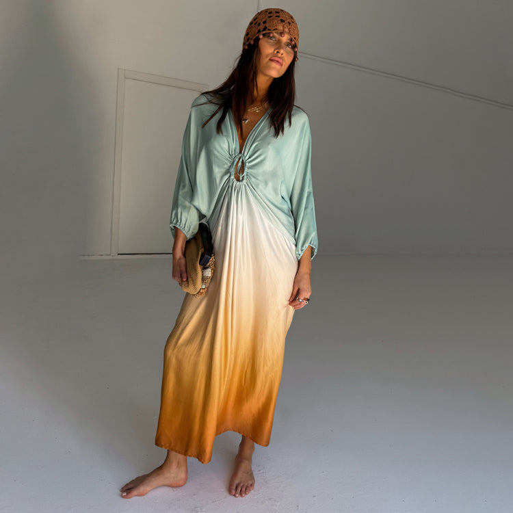 A woman wearing a blue ombre maxi dress with a tie bust detail, long sleeves, and a gradient from blue to orange.