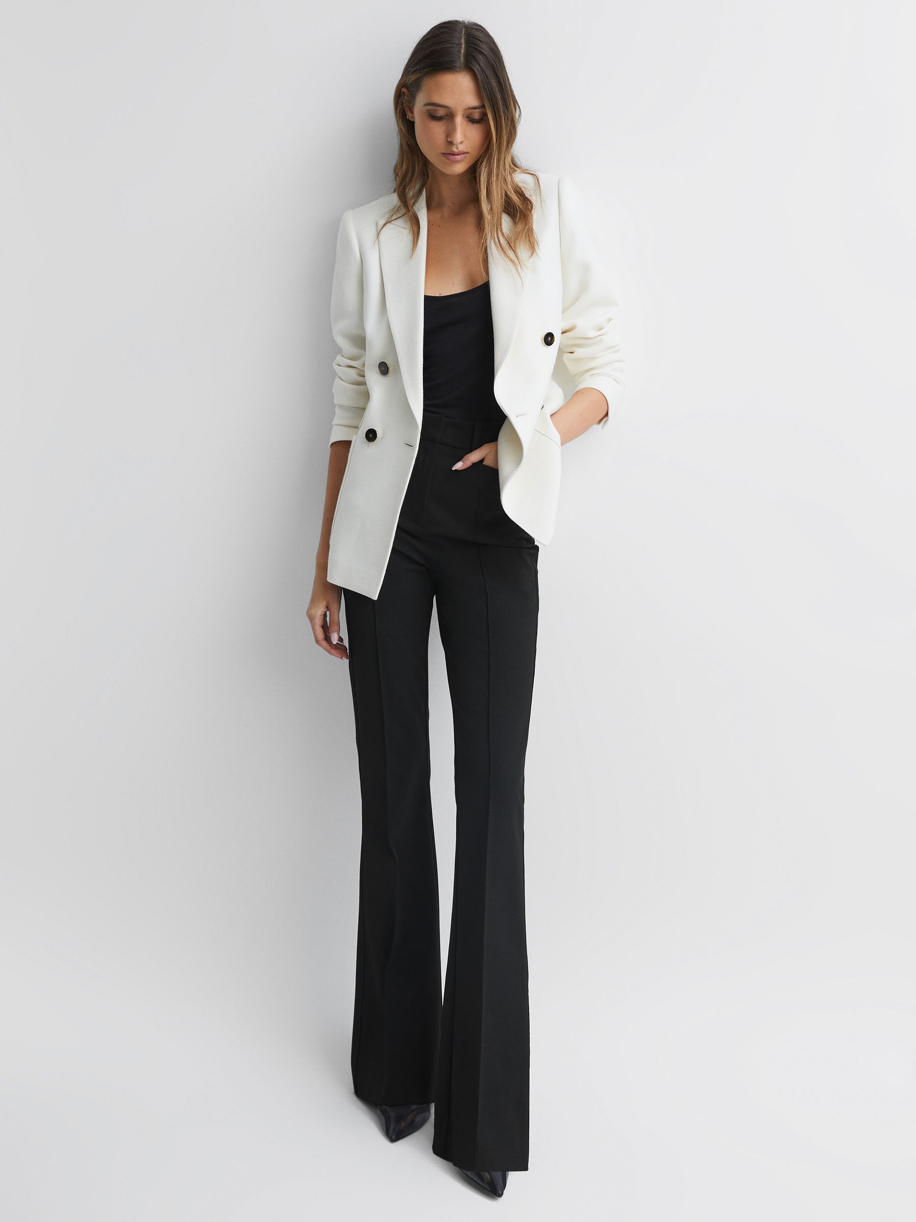 A woman wearing black flared high rise trousers with a white blazer and black top, standing against a white background.