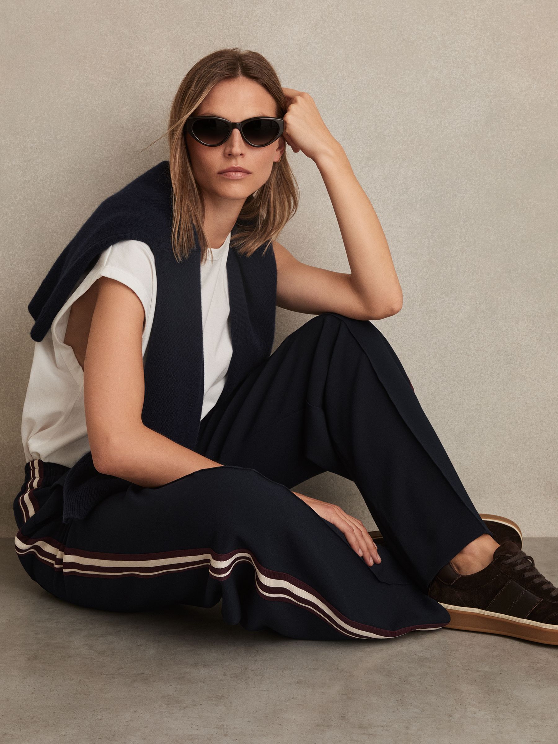 A woman wearing navy wide-leg trousers with side stripes, a white top, black sneakers, and sunglasses, sitting against a beige background.