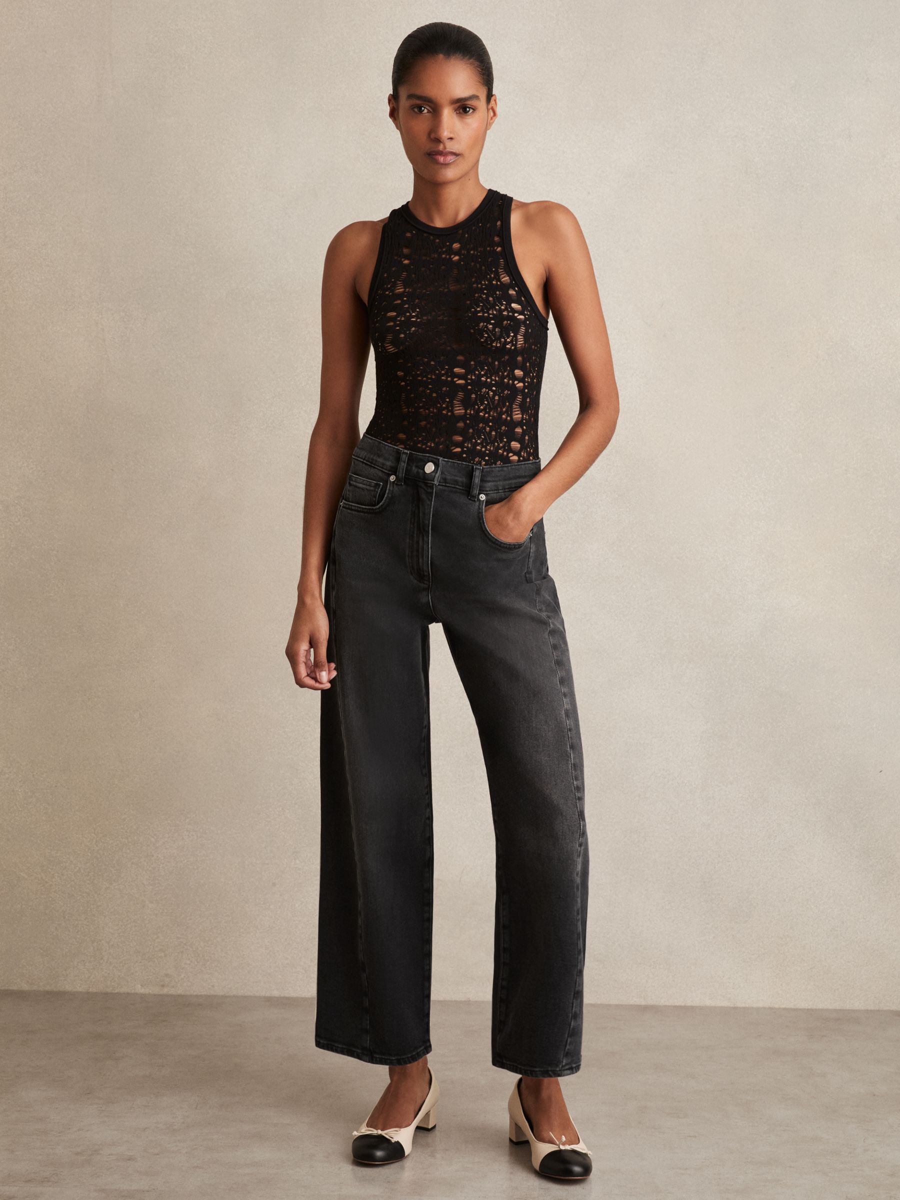 A woman wearing black jeans with seam details and a washed grey finish, paired with a black lace sleeveless top and beige heels.