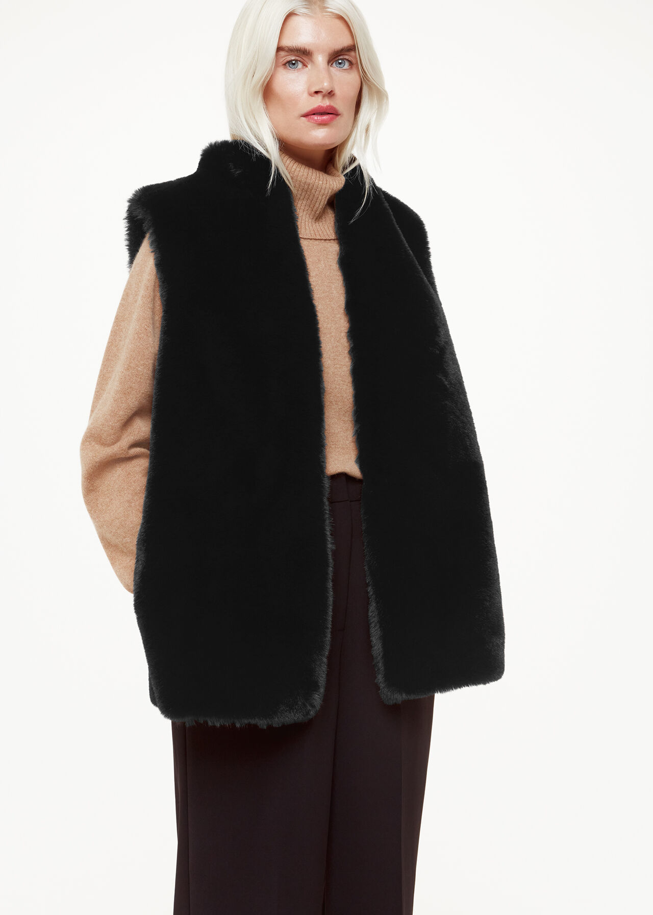 A woman wearing a black faux fur gilet with a high neck collar, sleeveless, mid-length, relaxed fit, over a beige sweater.