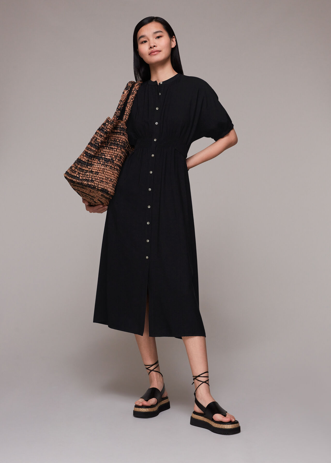 A woman wearing a black midi dress with a button-down front, tie belt, and two pockets, paired with black sandals and carrying a large woven bag.