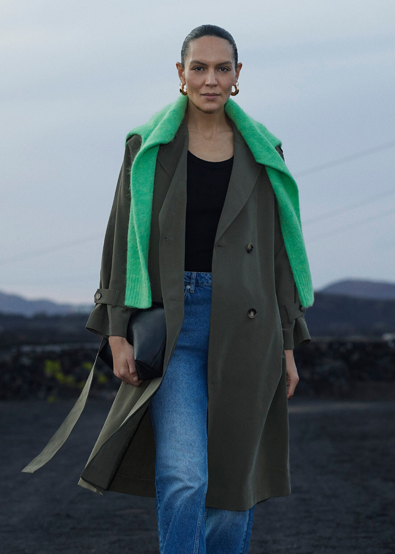 A woman wearing a khaki trench coat with a tie front, patch pockets, and a relaxed fit, standing outdoors.