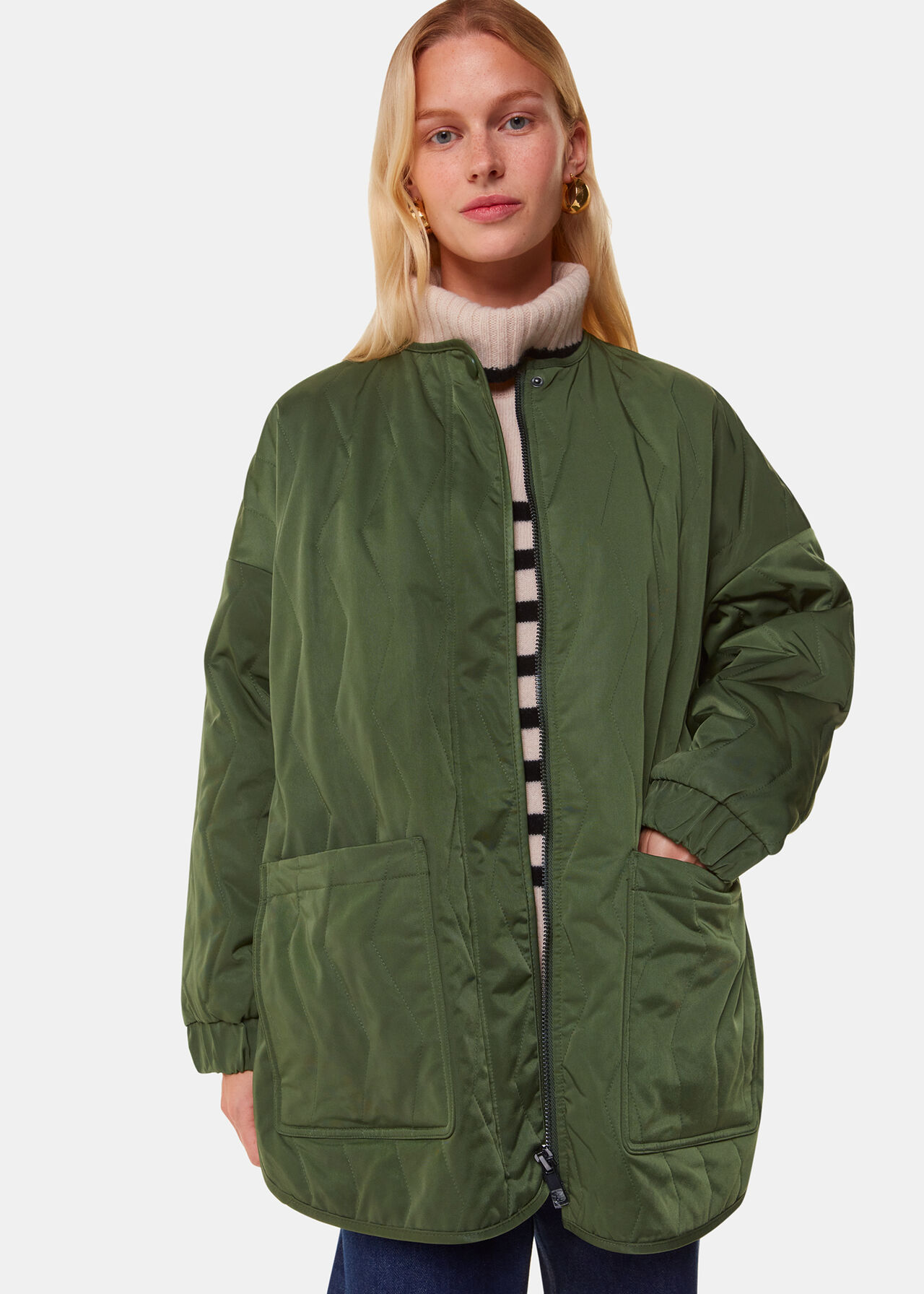 A woman wearing a khaki quilted coat with a cocoon silhouette, zig-zag stitching, large patch pockets, and a front zip with a popper button.