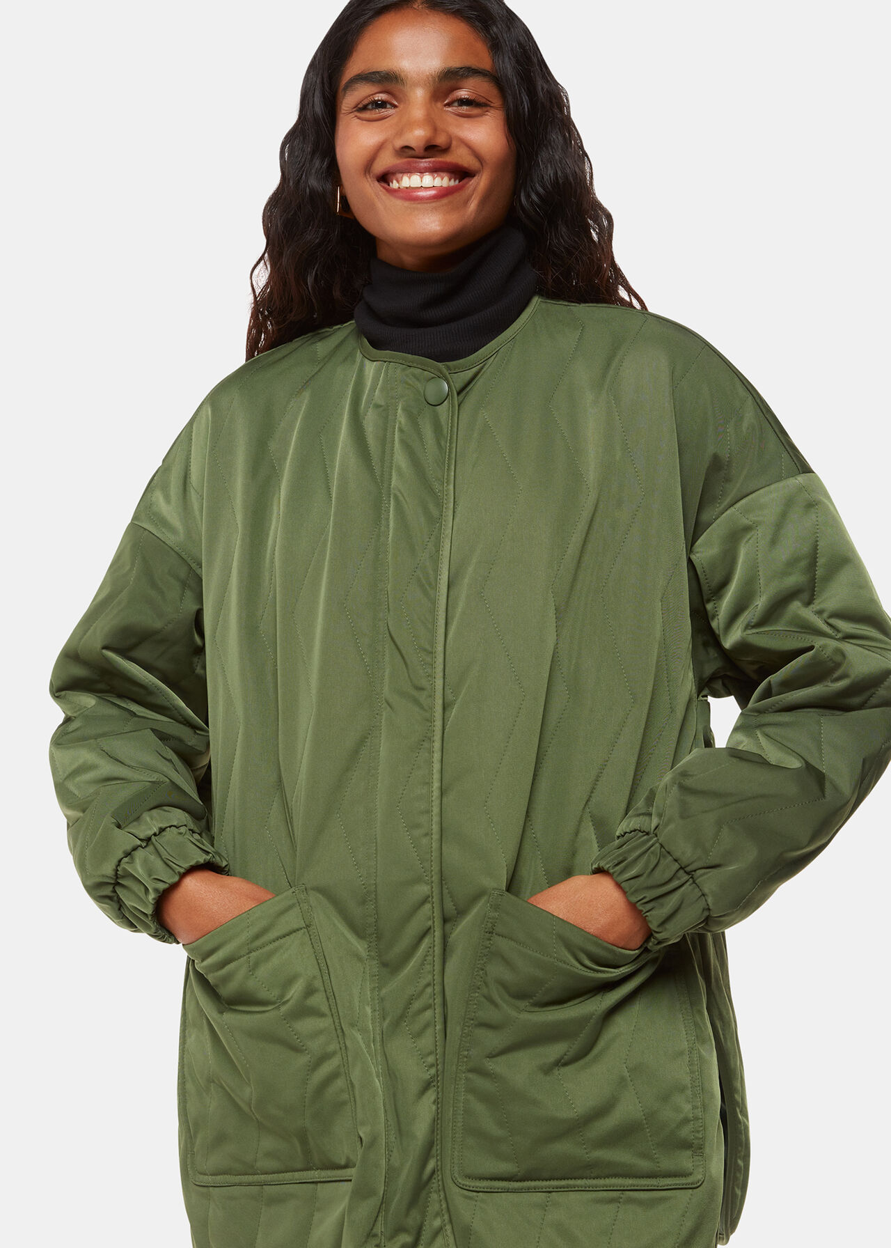 A woman wearing a khaki quilted coat with zig zag detailing, long sleeves, and large side pockets, with a zip and popper button closure.