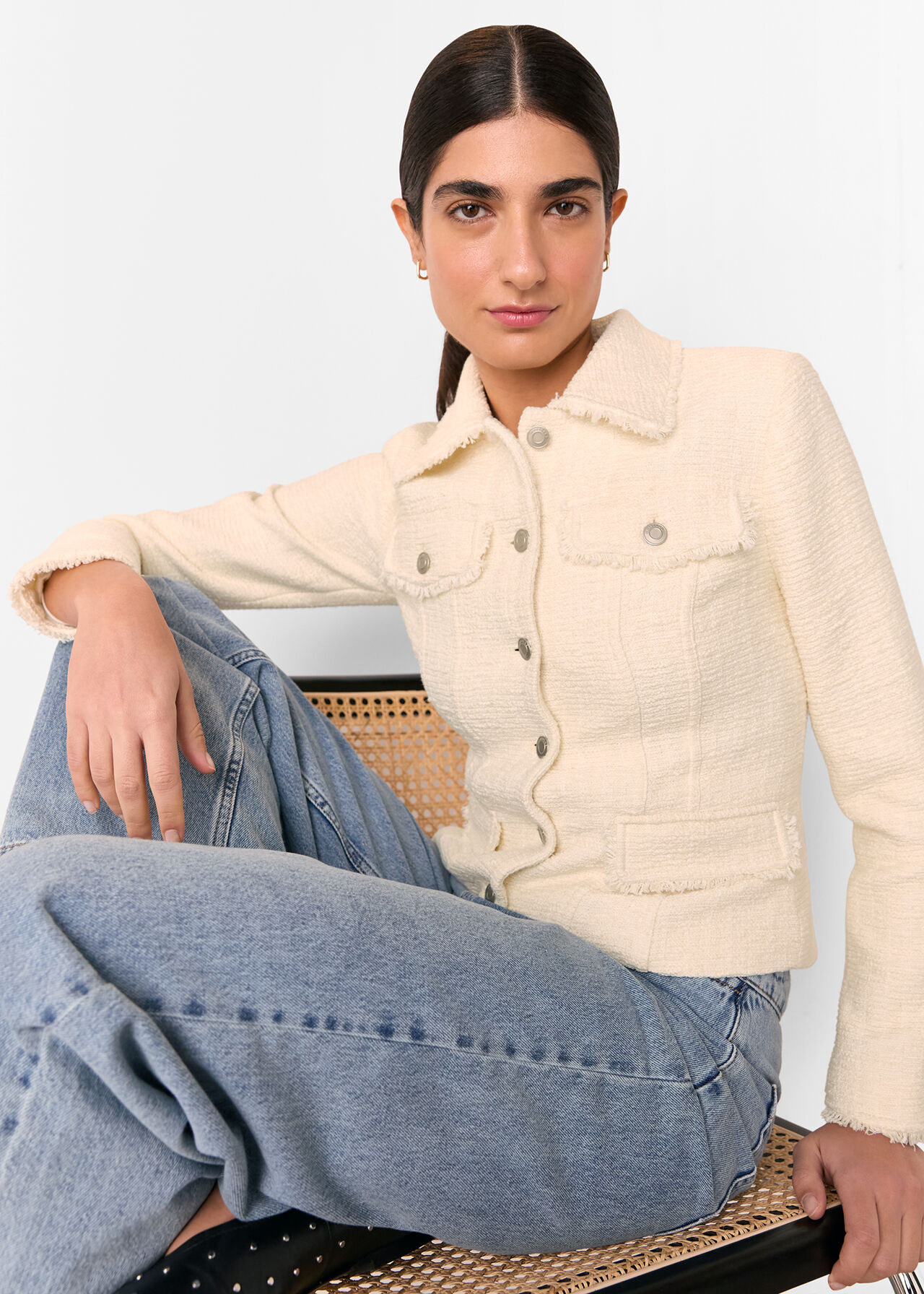 A woman wearing an ivory boucle jacket with denim buttons and frayed accents, sitting on a chair.
