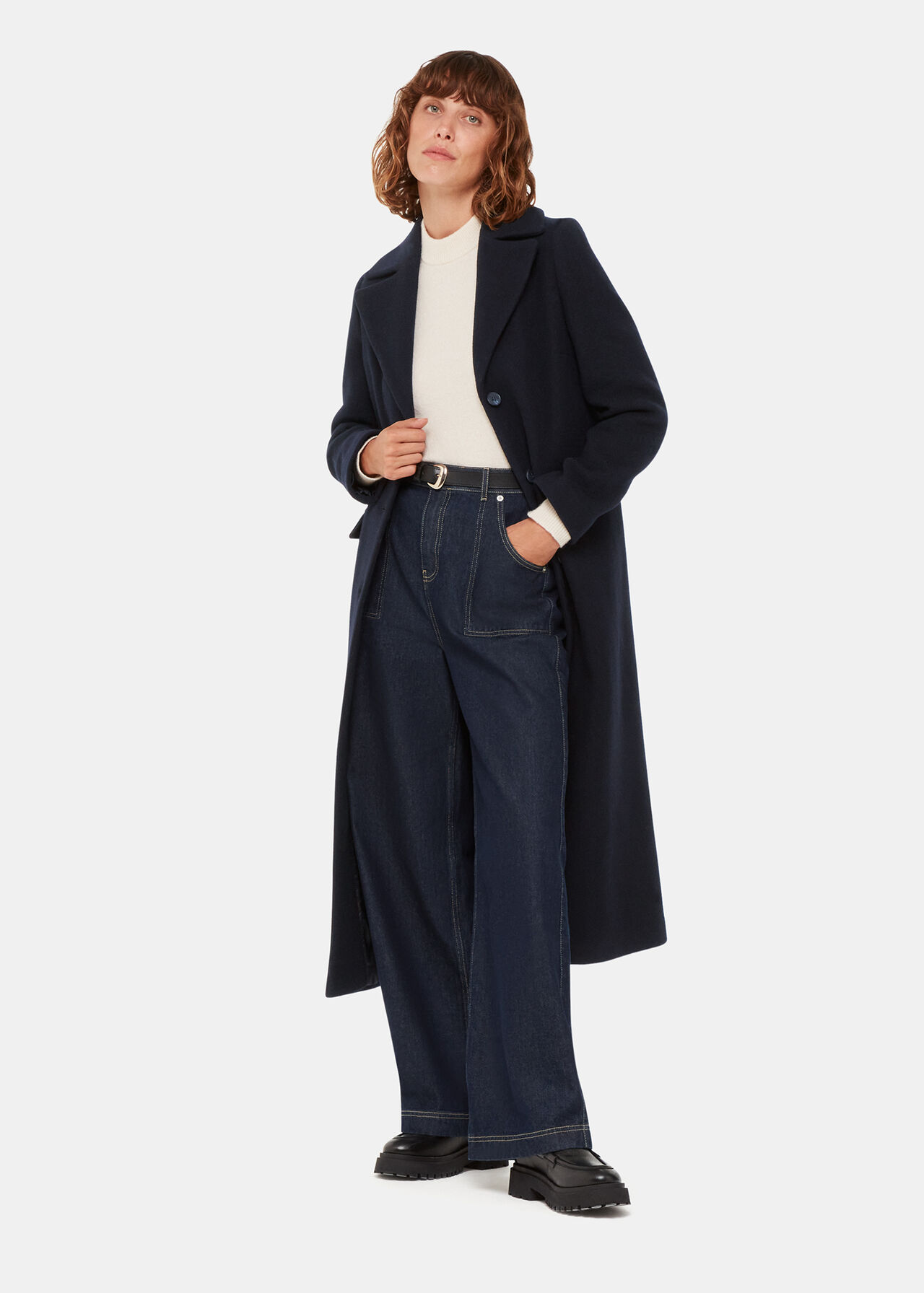 A woman wearing a navy longline coat with a notched lapel collar, three-button fastening, and two front pockets, paired with a white turtleneck and dark jeans.